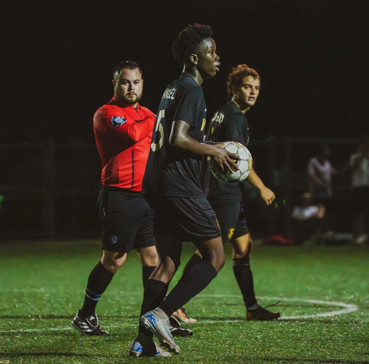 Soccer players on the field at night, with one holding a soccer ball, wearing black uniforms, and others in red and black uniforms.