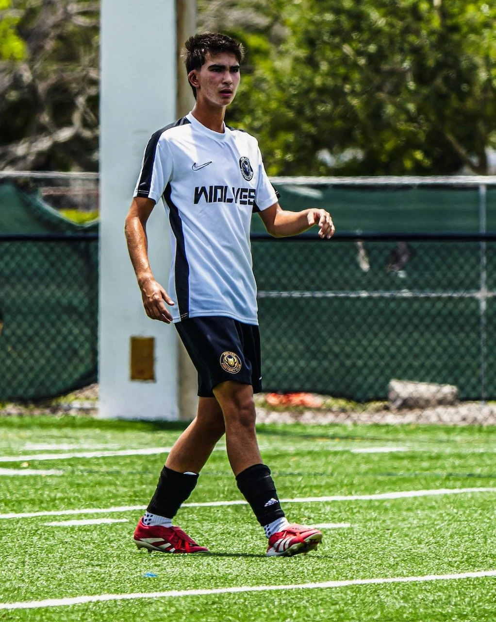 A young male soccer player with dark hair on a green field, wearing a white jersey with black accents and the word 'WOLVES' on it, black shorts, black socks, and red cleats.