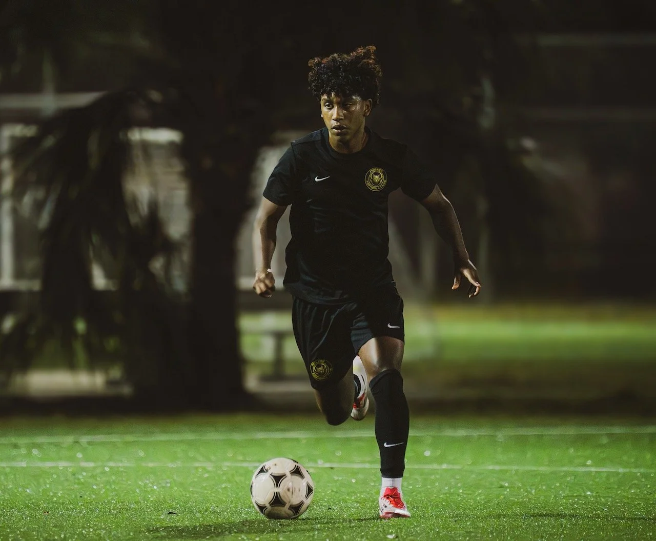 A young male soccer player in a black jersey and shorts is running on a green field, about to kick a soccer ball during night time.