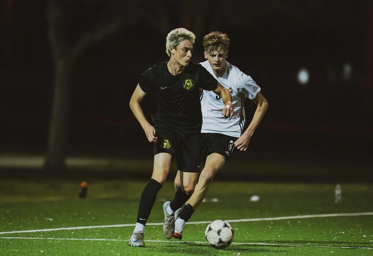 Two young male soccer players compete for the ball on a green field at night, illuminated by field lights.