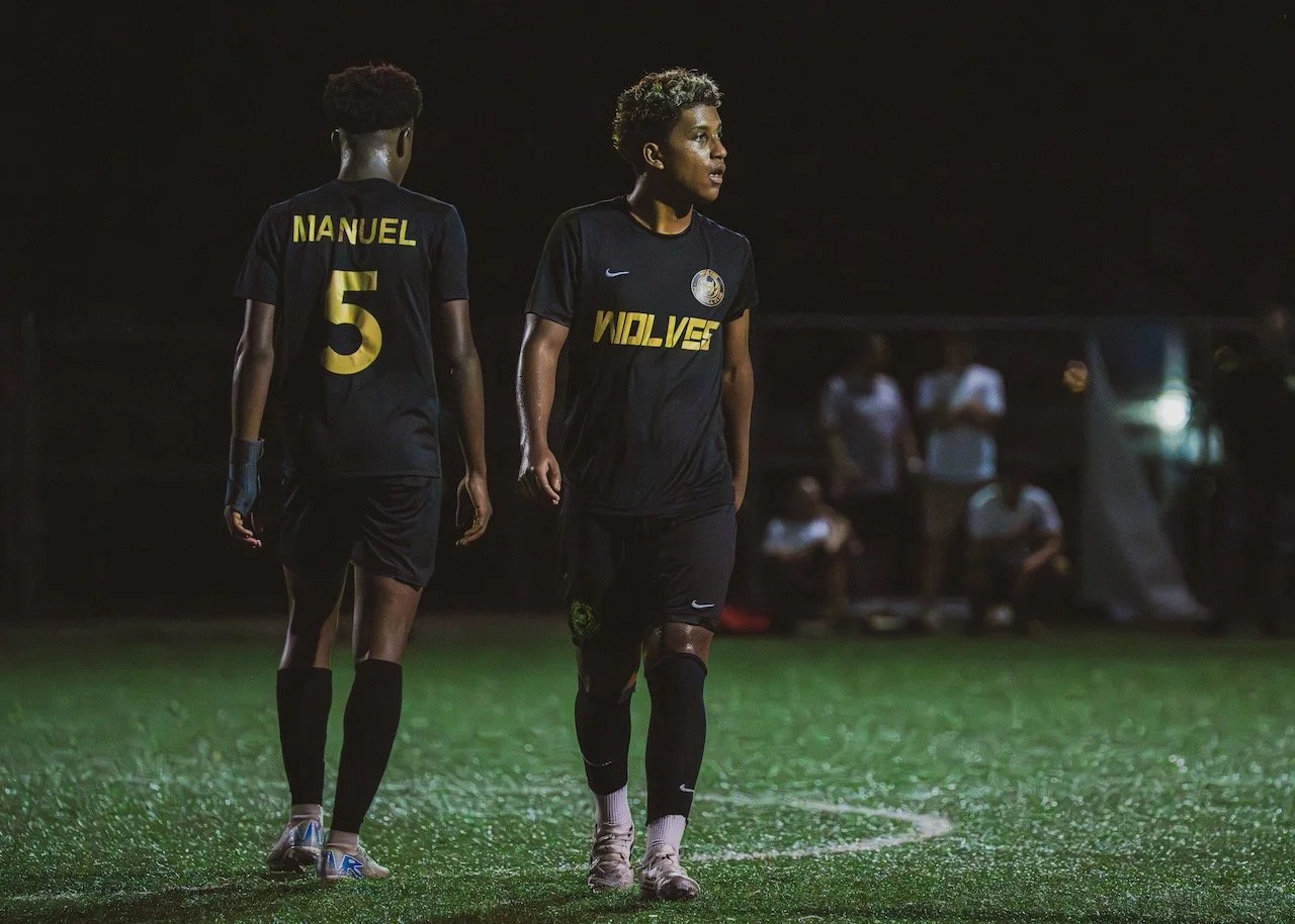 Two female soccer players in black uniforms walking on a field at night, with spectators in the background.