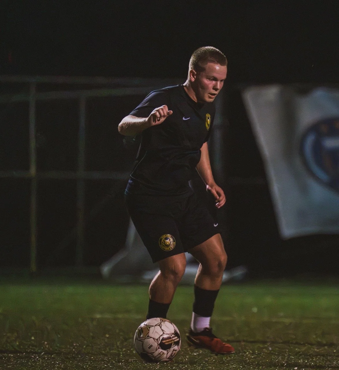 A male soccer player in black uniform with a yellow emblem dribbling a soccer ball on a field at night.