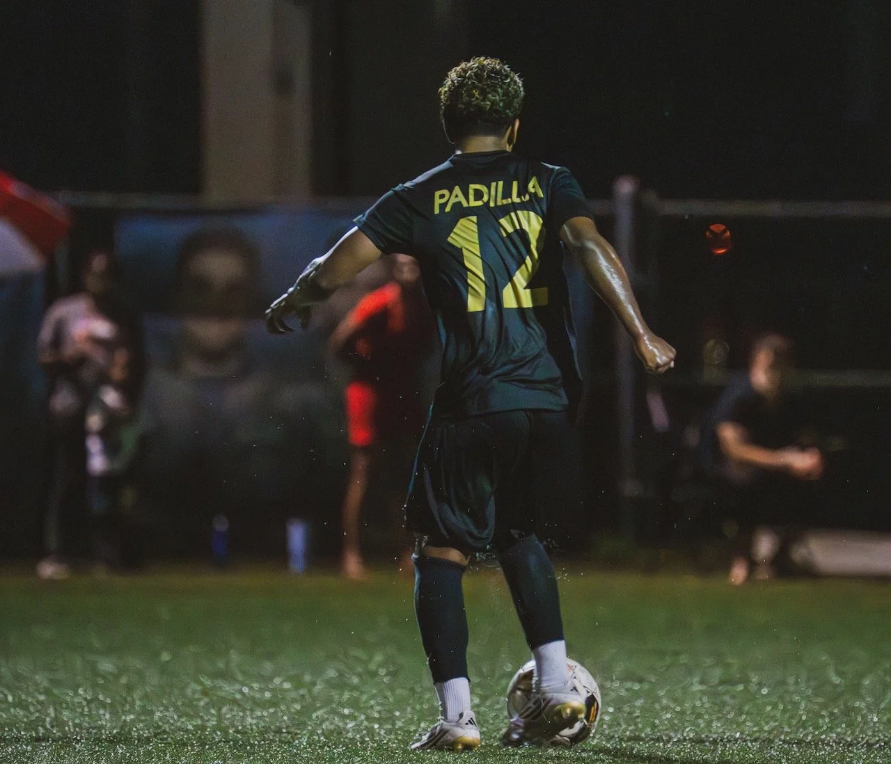 A soccer player wearing a black uniform with the name 'PADILLA' and number '12' on the back, contact with a soccer ball on a wet field at night.