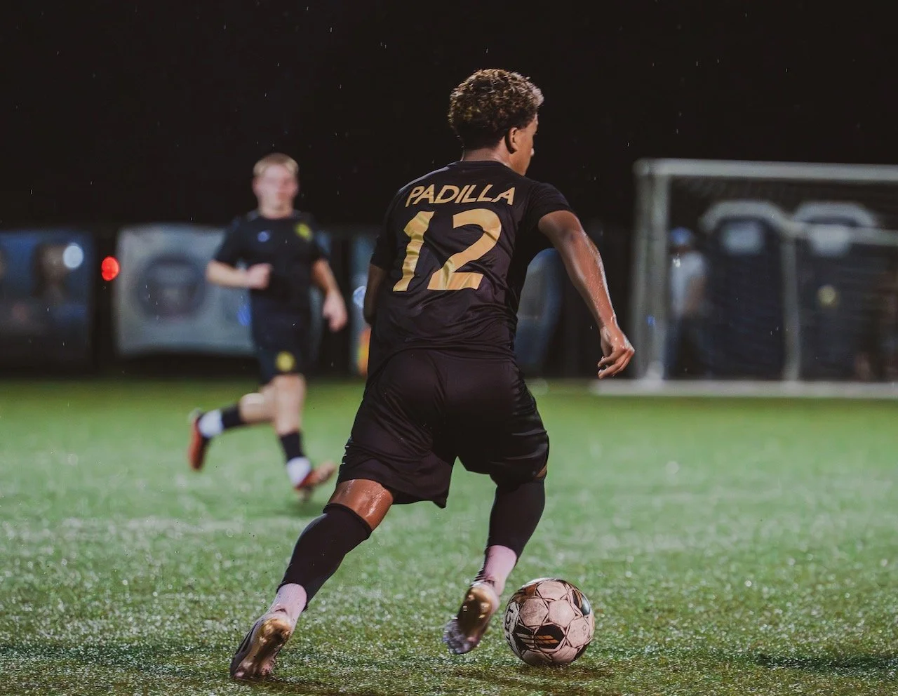 Soccer player with jersey number 12 kicking a ball on wet field at night, with another player in background.