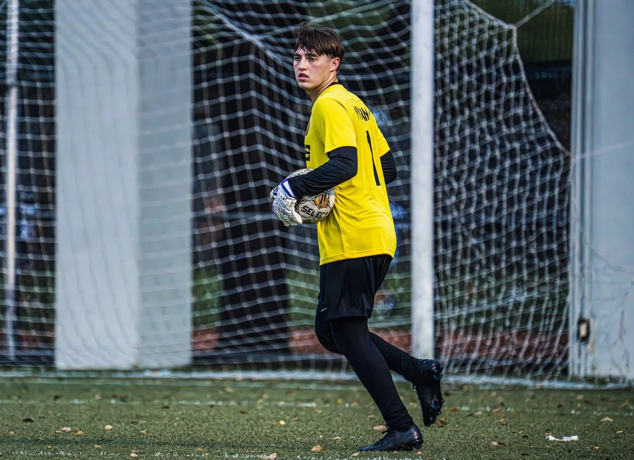 A young male soccer goalkeeper in a yellow jersey and black athletic gear standing on a soccer field, holding a soccer ball with a netted goal in the background.