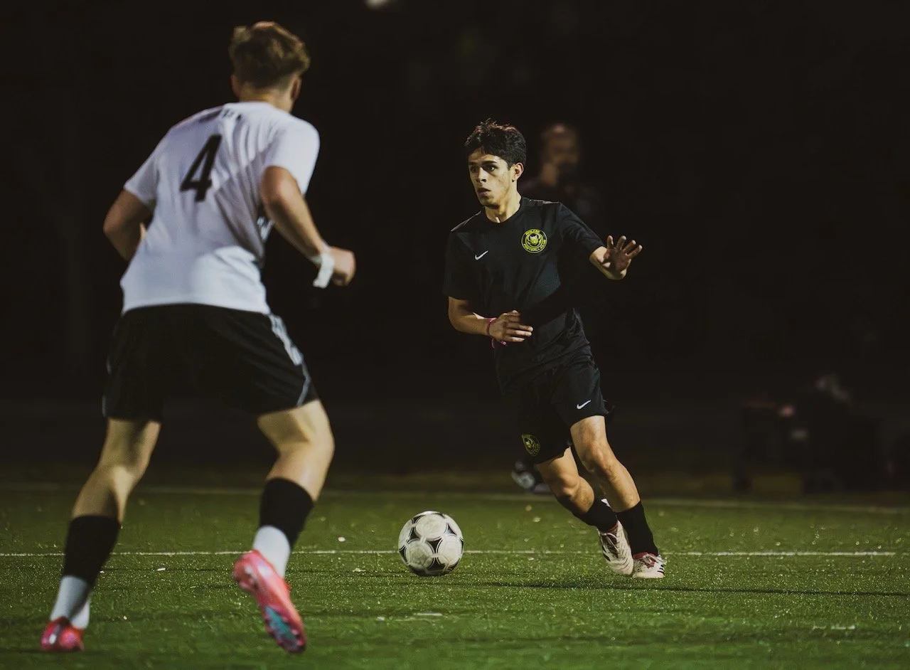 Two young male soccer players competing for the ball on a field at night. One player is dressed in a black uniform, and the other in a white jersey with the number 4. The player in black is approaching the ball while the player in white is defending.