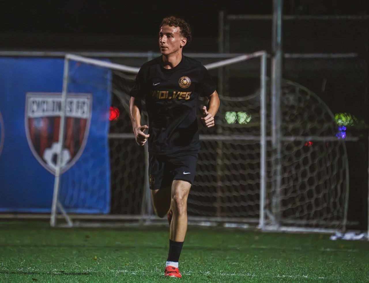 A young soccer player running on the field at night, wearing a black uniform with gold and white details, near goalpost with a Wolves logo seen on the jersey.
