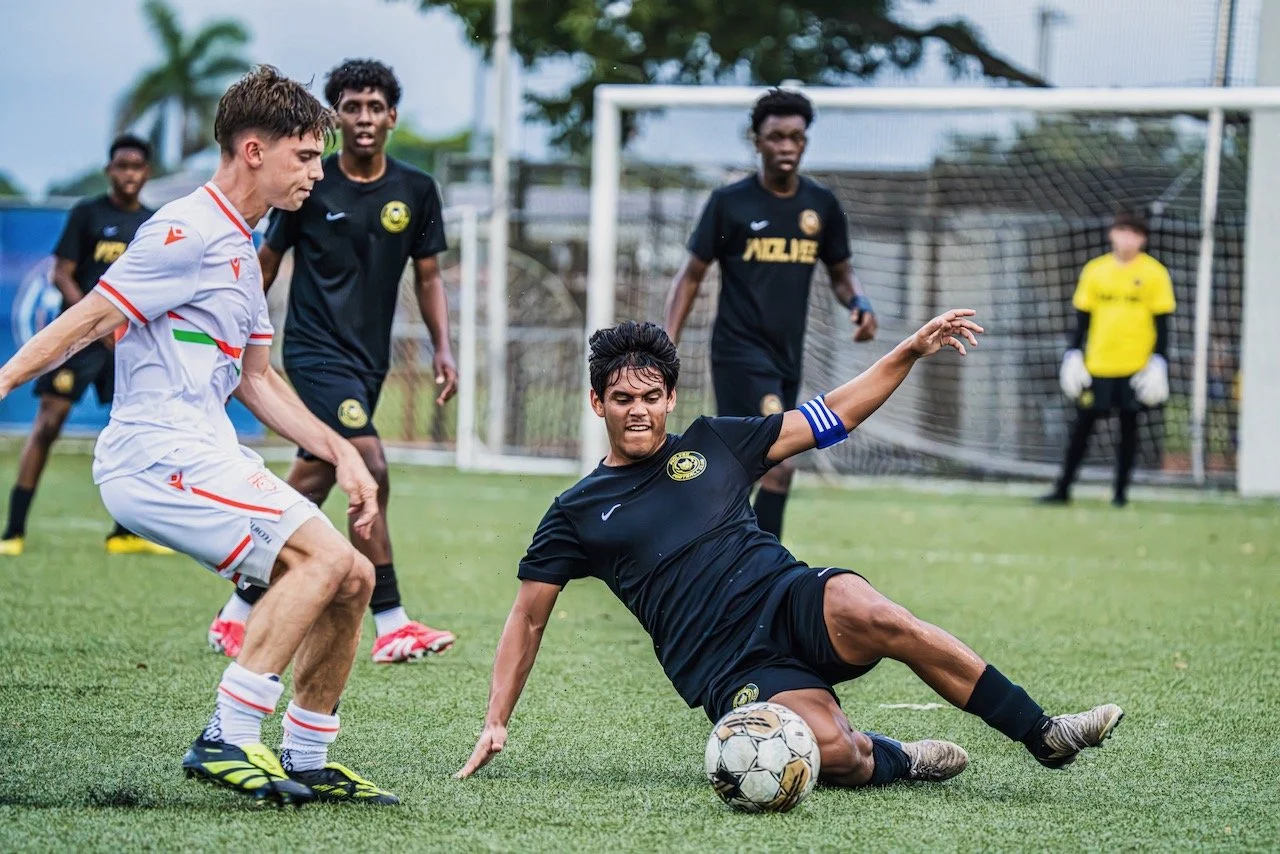 Soccer match where a player in black is sliding on the ground, attempting to control the ball, while an opponent in white tries to challenge him. In the background, other players and a goalkeeper in yellow are visible near the goal.