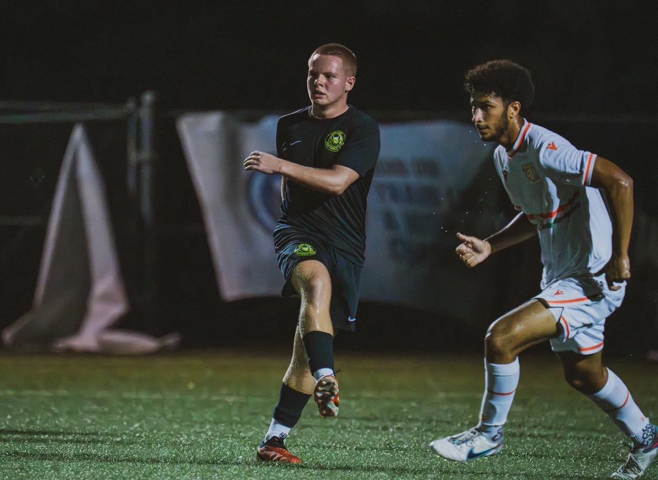 Two soccer players competing for the ball on a rainy night field, one in black uniform and one in white uniform.