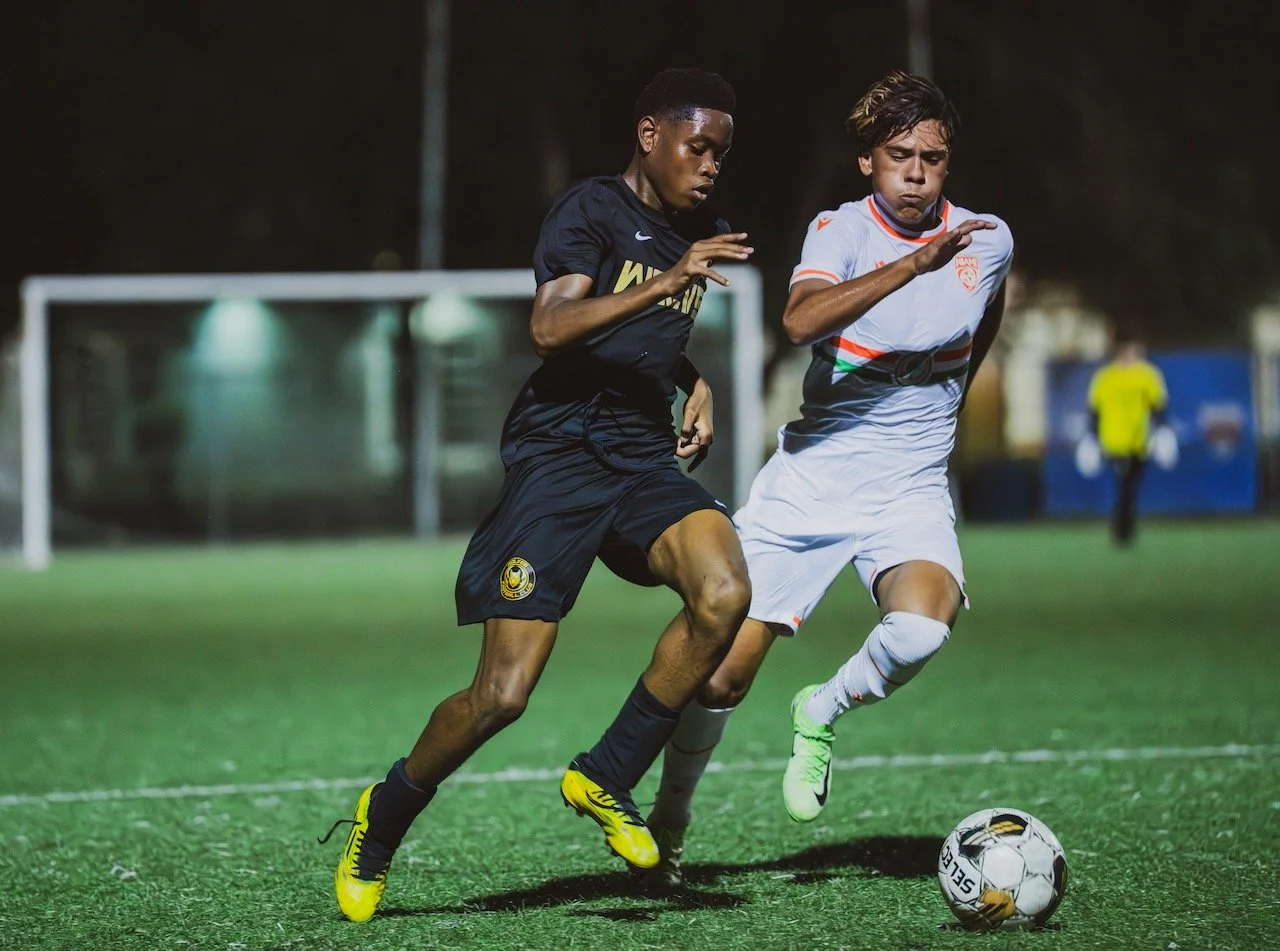 Two soccer players competing for the ball on a green field during a match at night, with a goalpost and a goalkeeper in the background.