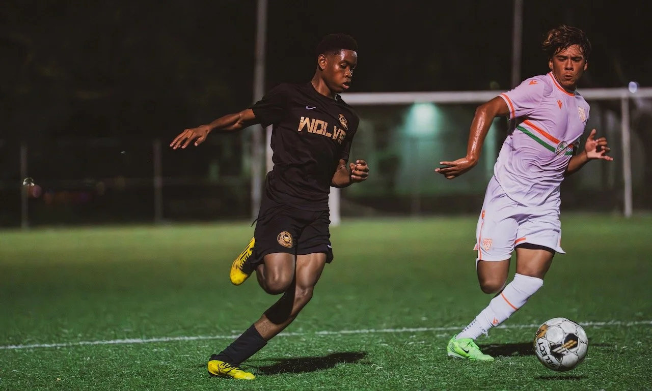 Two soccer players chasing a ball on the field at night, one wearing a black uniform and the other wearing a white uniform with orange and green accents.