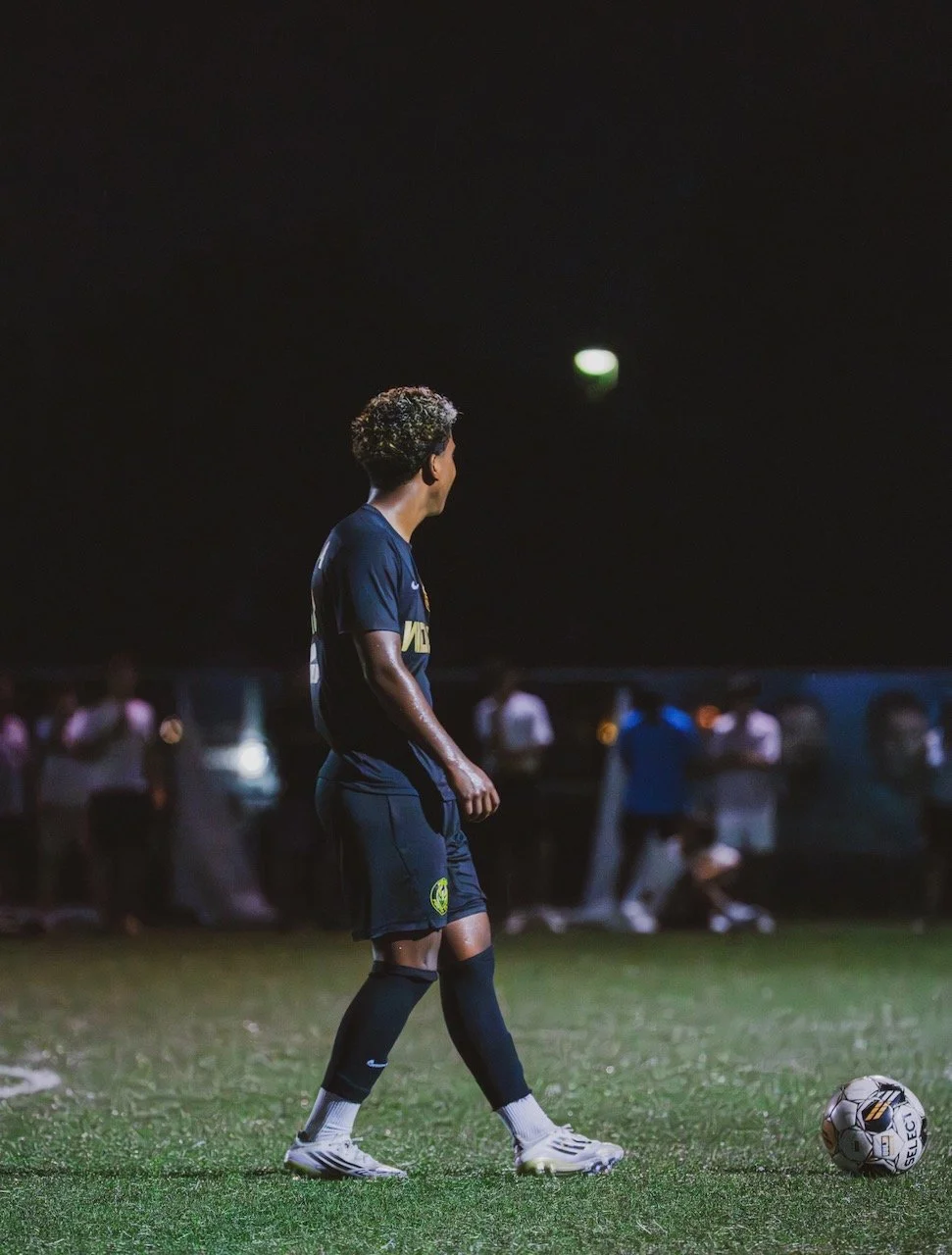 A male soccer player standing on a well-lit grass field at night, about to kick a soccer ball, with people watching in the background.