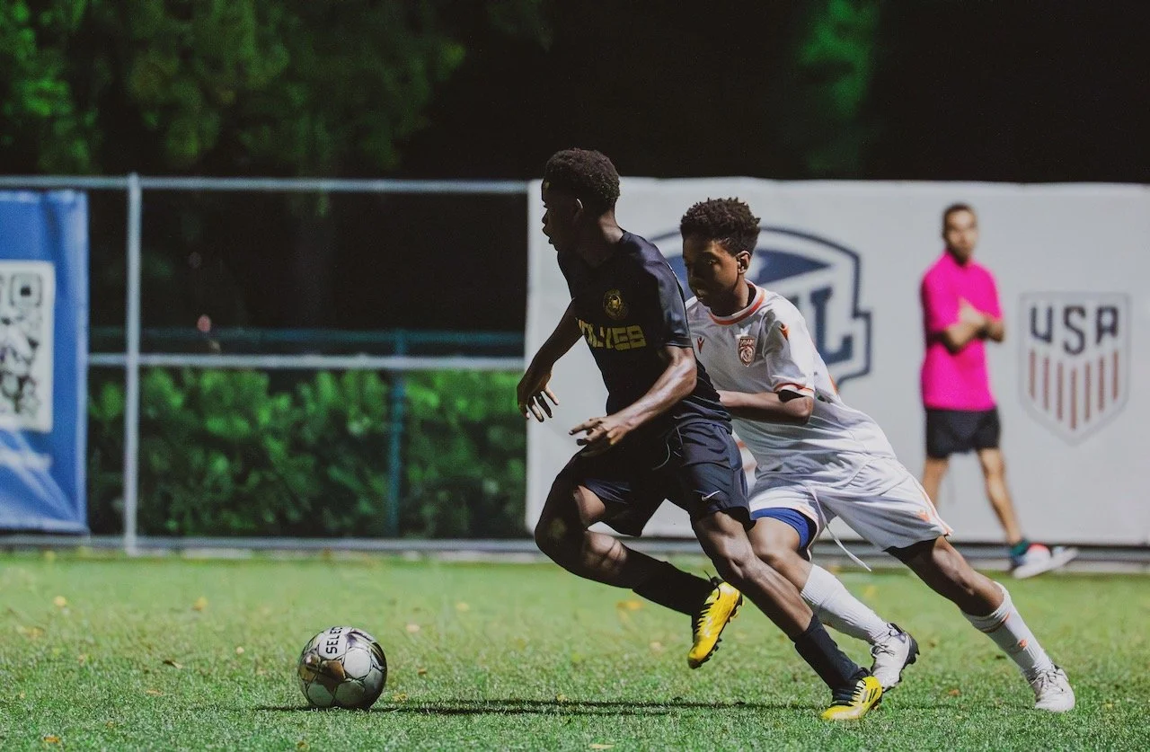 Two young soccer players racing for the ball on a field during a game, with a USA team official or referee watching in the background.