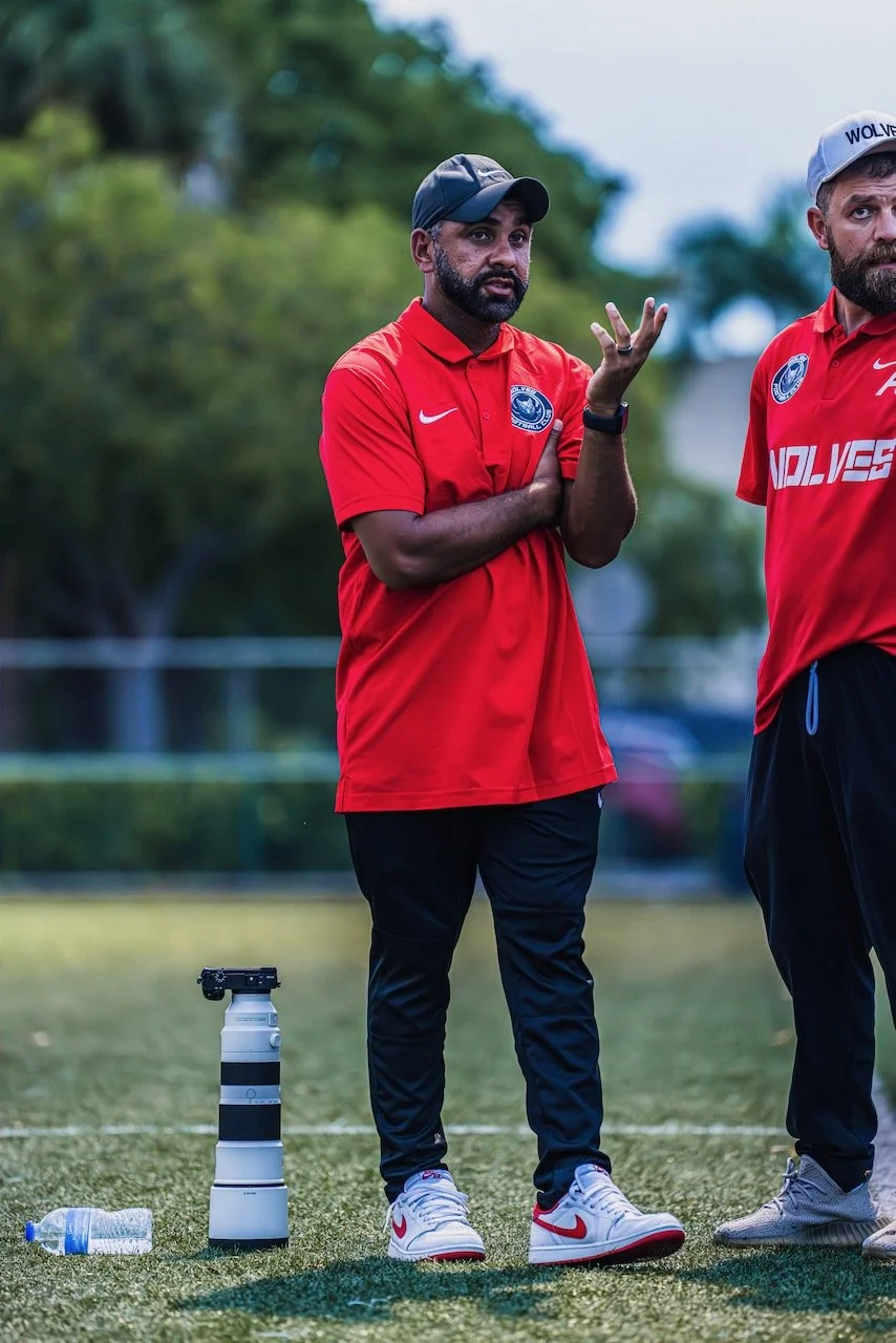 Two men in red sports jerseys standing outdoors on a field, one appears to be explaining or gesturing, with trees and a water bottle on the ground nearby.