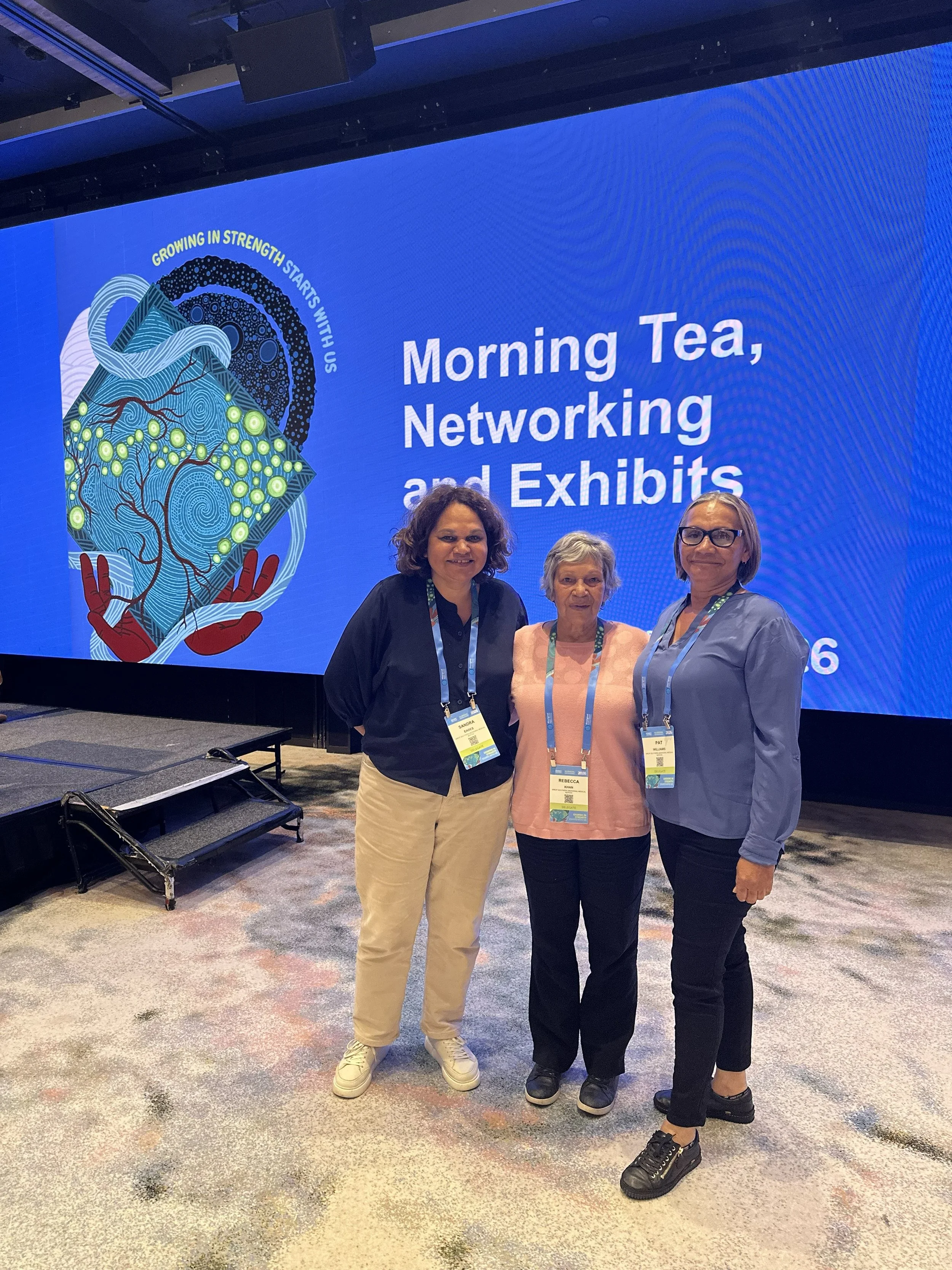 Three women standing together at a conference, smiling in front of a large blue screen that reads 'Morning Tea, Networking and Exhibits,' with artwork of a mountain, a tree, and abstract patterns. They are wearing conference badges.