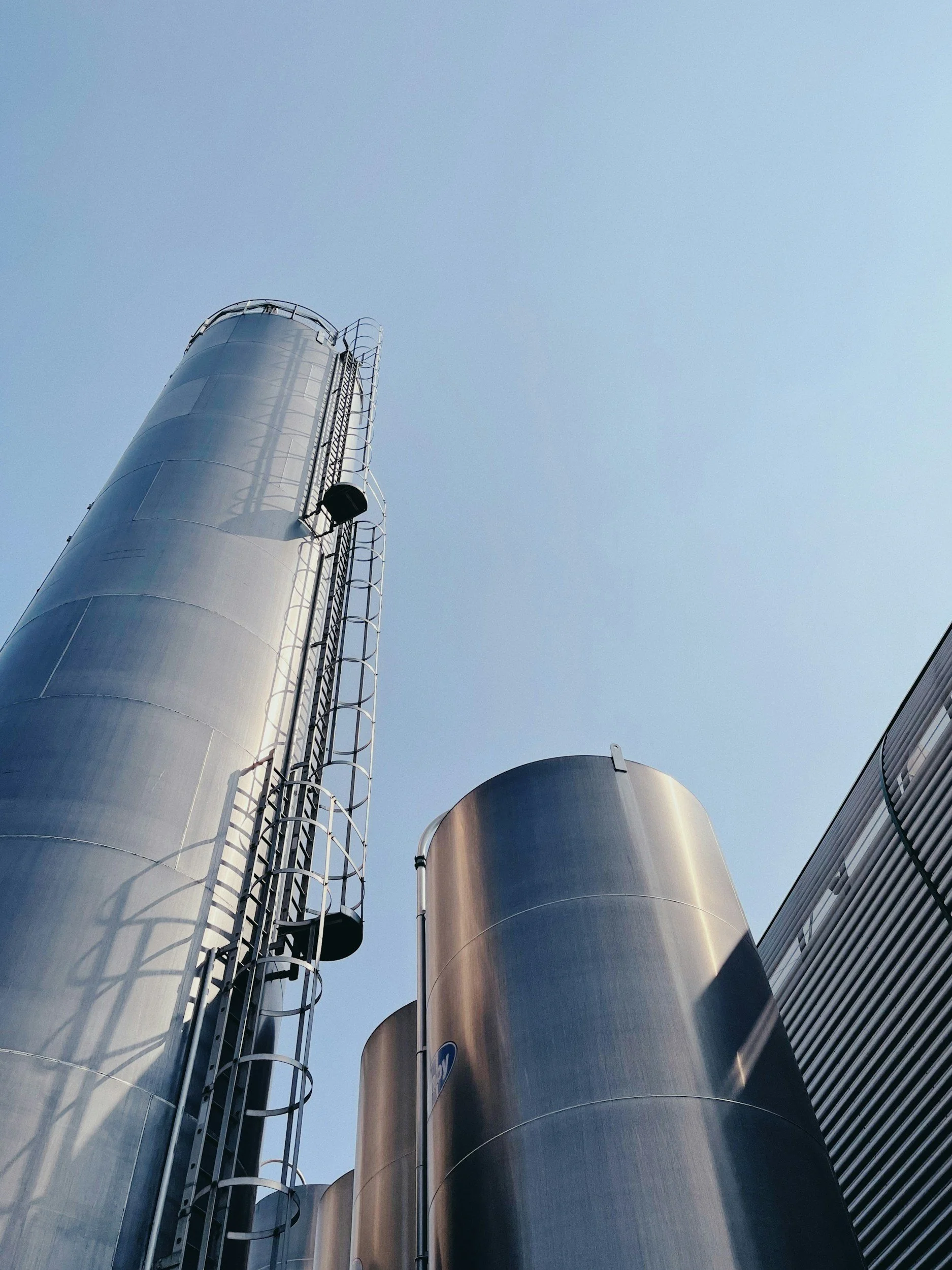 Looking up at tall cylindrical industrial silos made of metal, with external ladders and railings, against a clear blue sky.