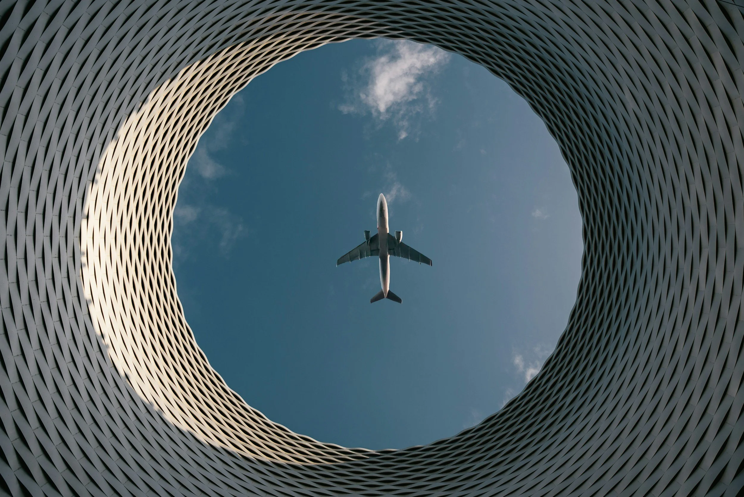 Looking up through an architectural structure with a circular opening at the top, an airplane flying in the sky is visible.