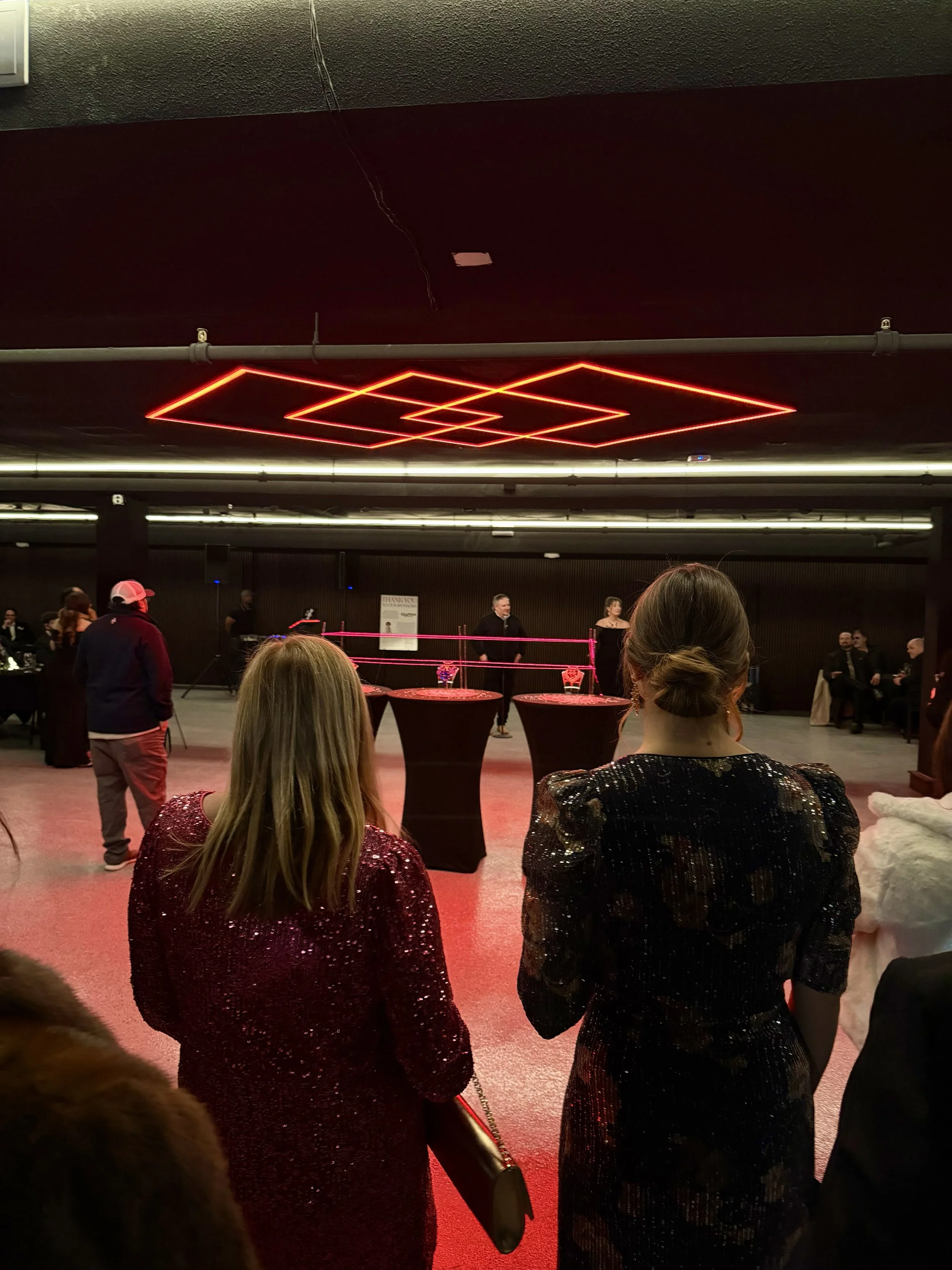 People attending a formal event in a dimly lit room with red lighting and decorative ceiling lights. Some are dressed in sequined clothing, and there are high tables in the room, suggesting a cocktail or reception setting.