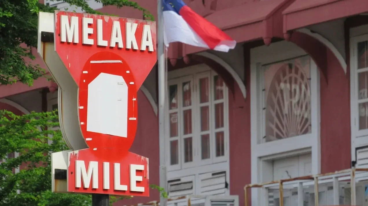Iconic red "MELAKA MILE" sign standing in front of the historic red Dutch colonial building (Stadthuys), with the Malaysian or Melaka flag flying above, marking travel to the UNESCO heritage city of Melaka, Malaysia.