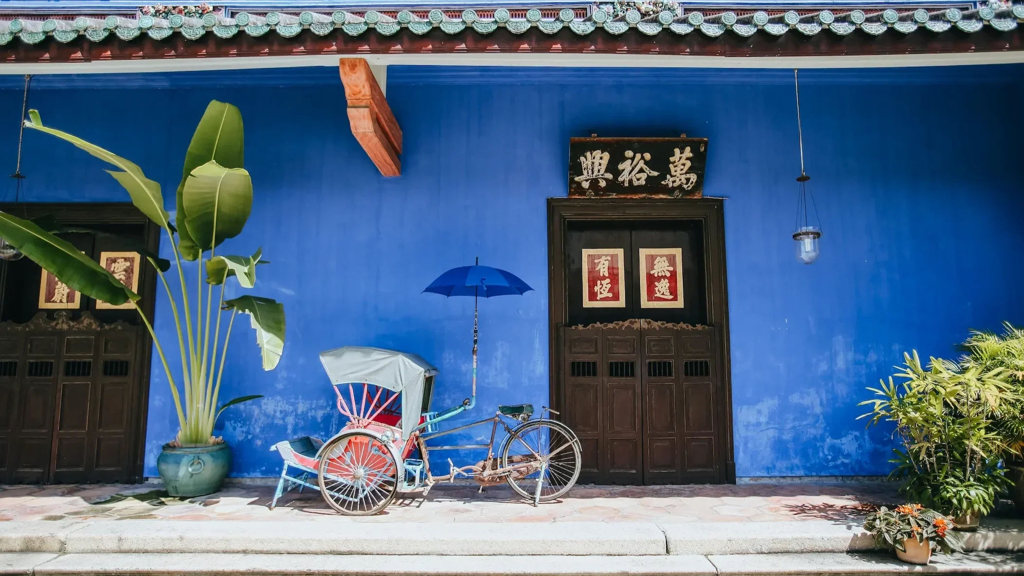 Traditional, decorated trishaw parked in front of an iconic blue heritage wall and a weathered Chinese store sign in Penang.