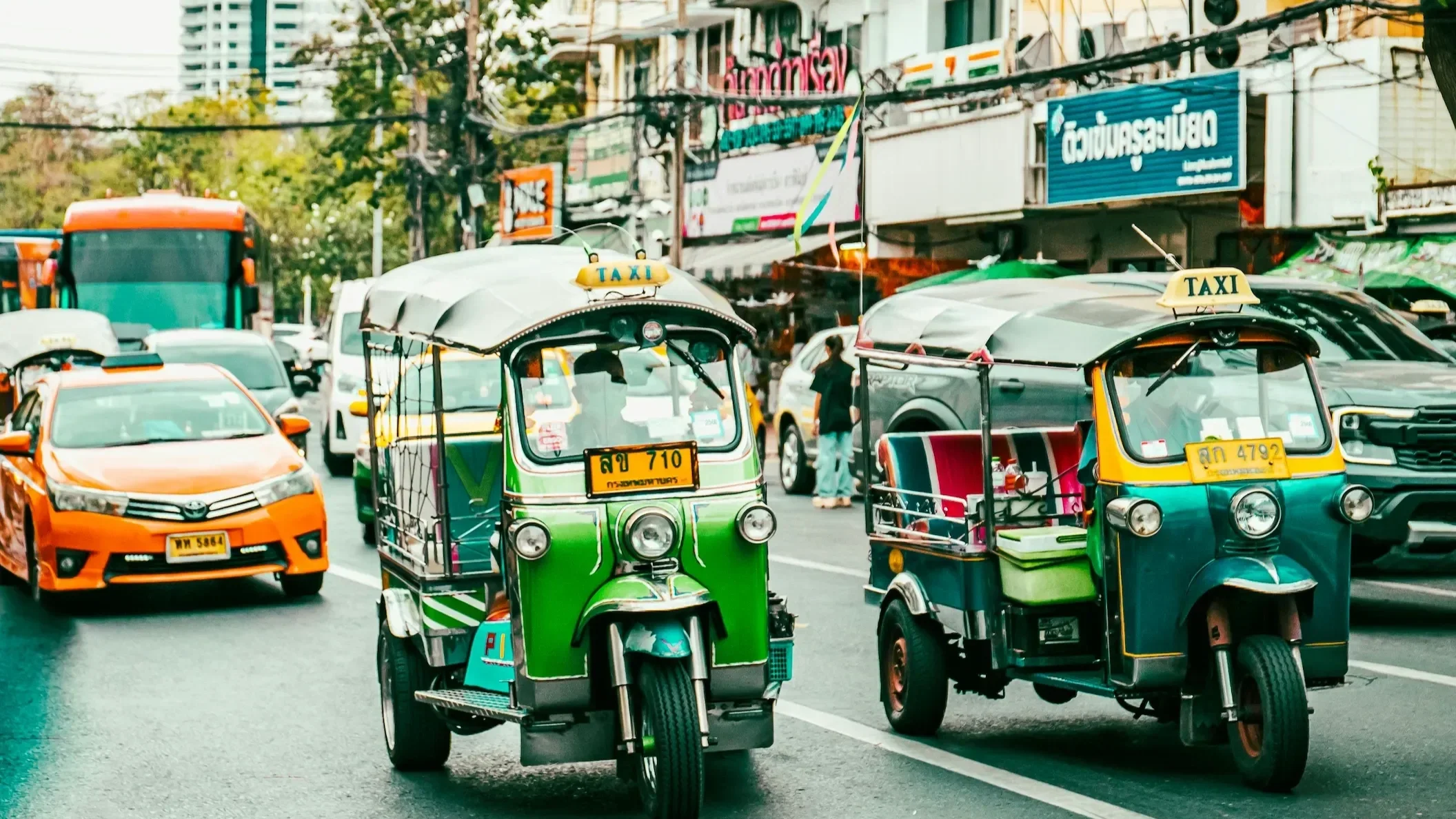 Two brightly colored, iconic Tuk-Tuk taxis (auto rickshaws) navigating a busy Bangkok street amidst traffic. The image represents thrilling urban travel and Southeast Asian adventure in Thailand.