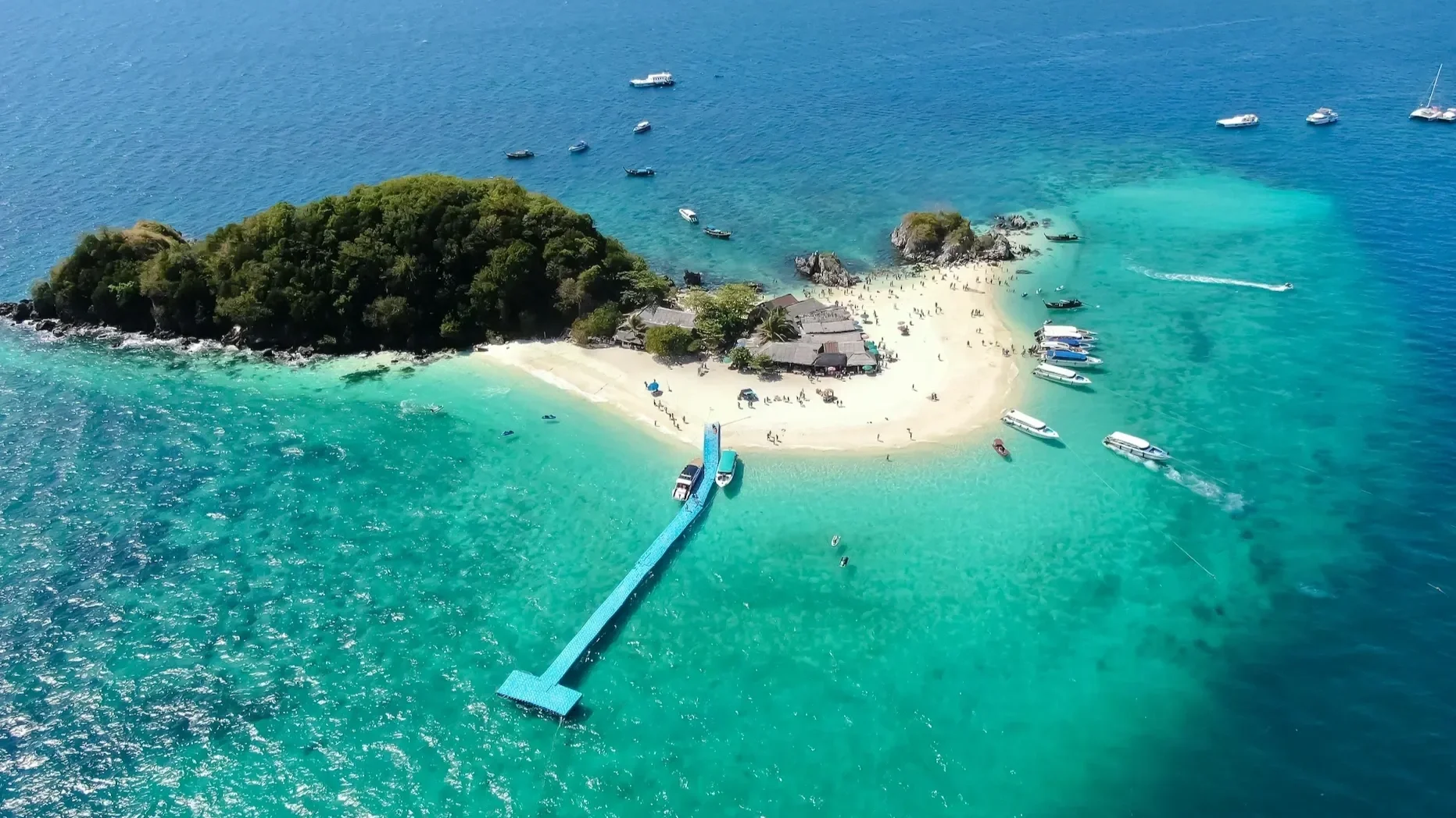 Aerial view of a small tropical island near Phuket, Thailand, featuring crystal clear turquoise water, a white sand beach, and a long blue jetty.