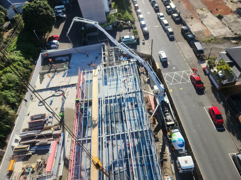 Construction workers working on building a structure with steel framing, with a concrete pump truck pouring concrete, next to a street with parked cars and nearby houses.