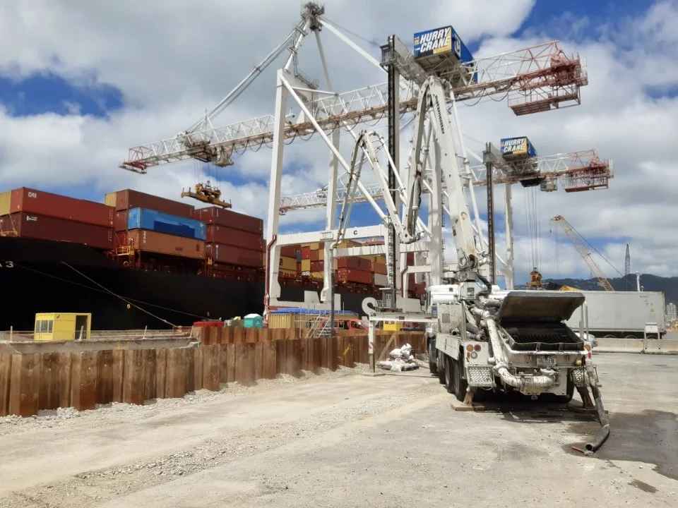 A large ship docked at a port filled with stacked shipping containers, with multiple tower cranes overhead, and a concrete pump truck in the foreground pouring concrete.