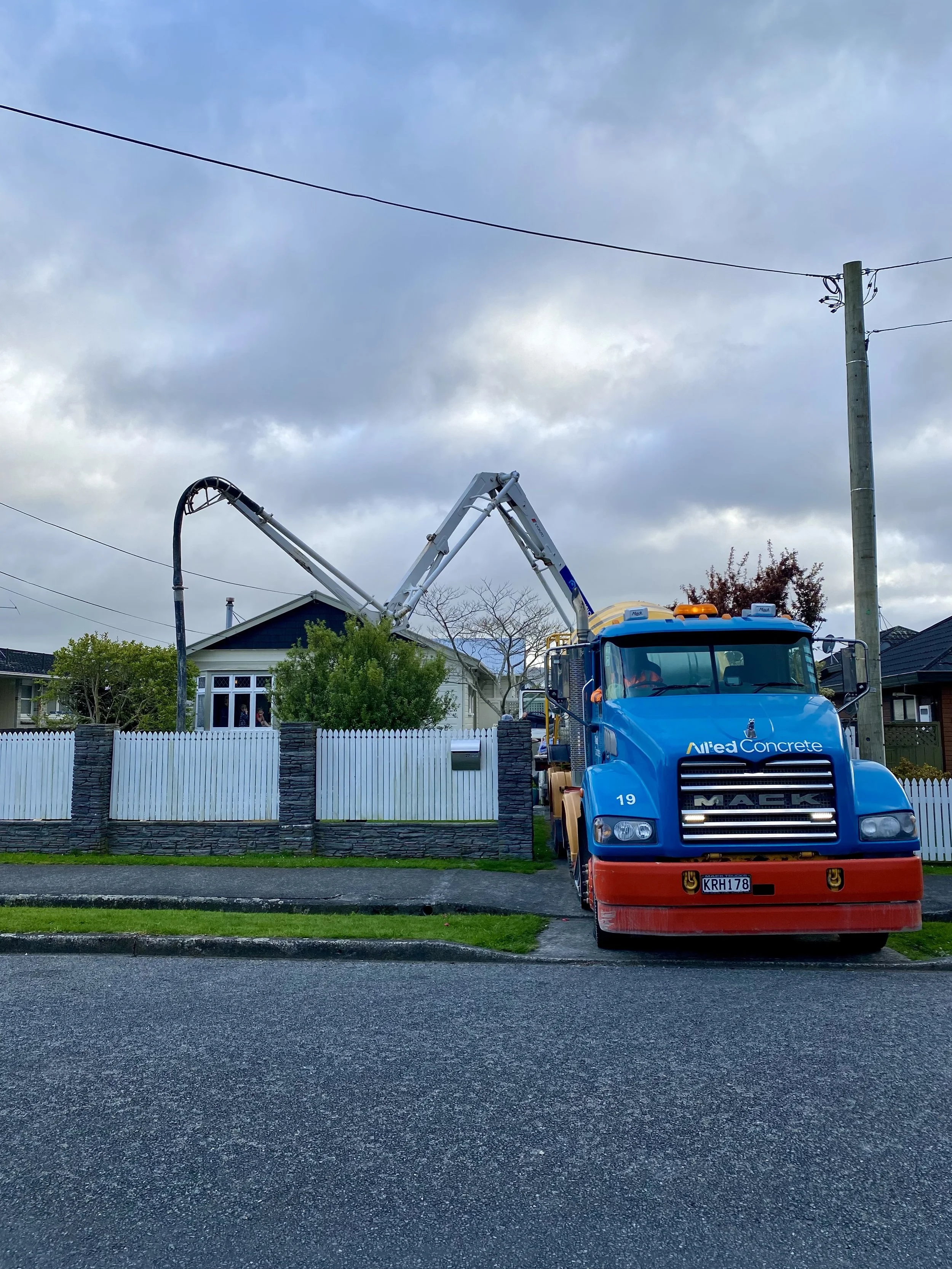 A large blue concrete pump truck parked on a residential street in front of houses with trees and a white fence, under a cloudy sky.