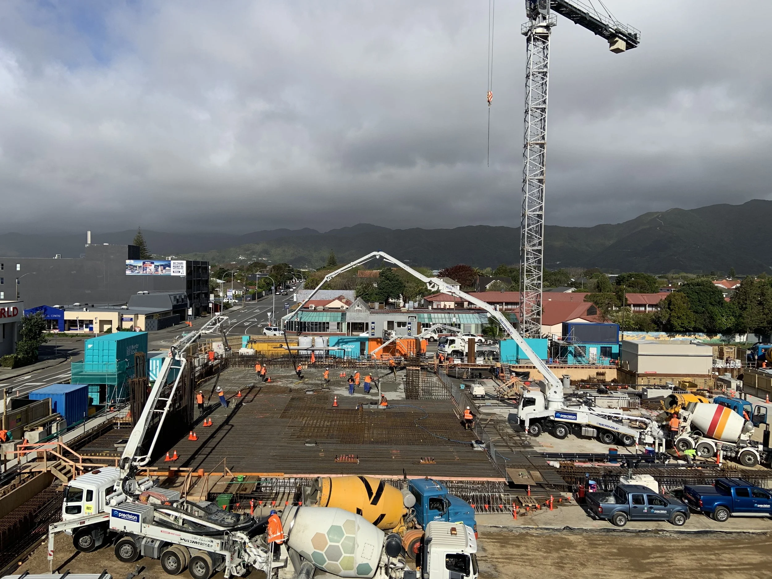 Construction site with workers, cement mixers, trucks, cranes, and unfinished building structure under cloudy sky.