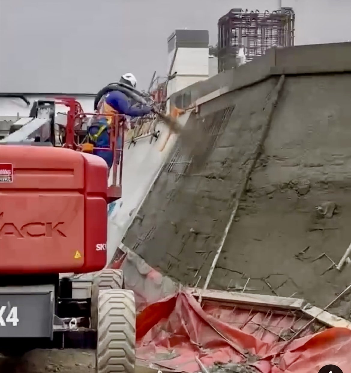 Construction worker wearing safety gear uses a hydraulic breaker attachment on a red excavator to break concrete on a building site under a cloudy sky.
