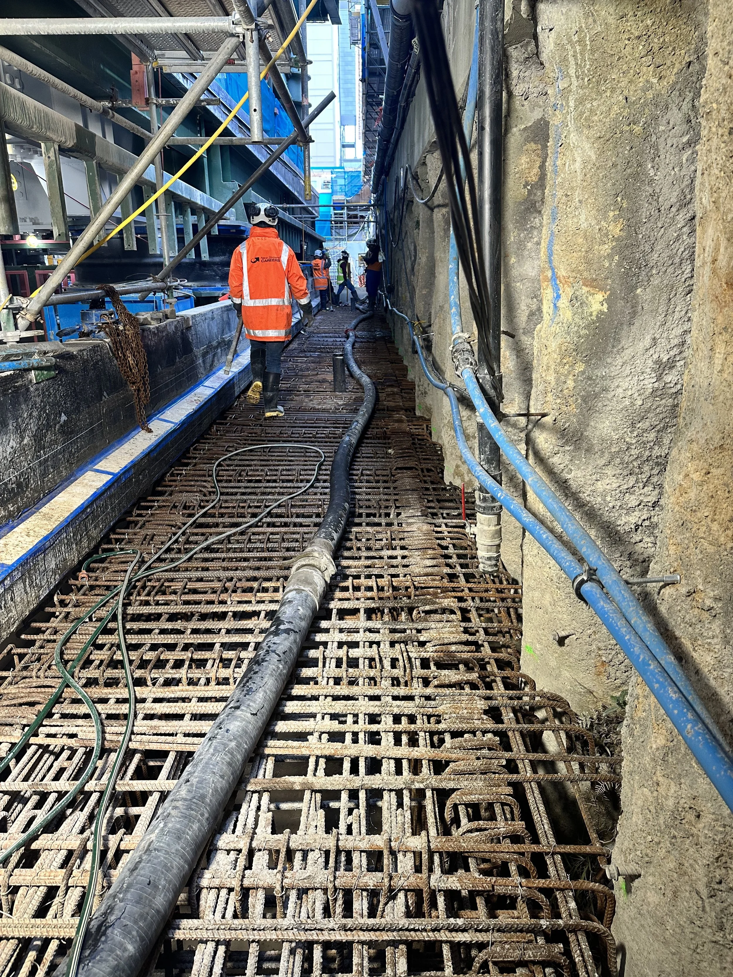 Construction workers in safety gear including helmets and high-visibility vests are working on a building site. They are walking on a steel rebar grid foundation with construction pipes and cables placed along the site. Scaffolding and a partially built structure are visible on the left, and a concrete wall with exposed rebar is on the right.