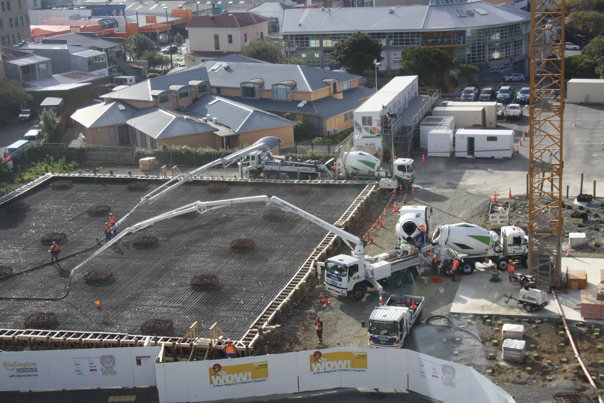 Construction site for a new children's hospital with workers, concrete mixers, and building frameworks; neighboring buildings and parking lot in the background.