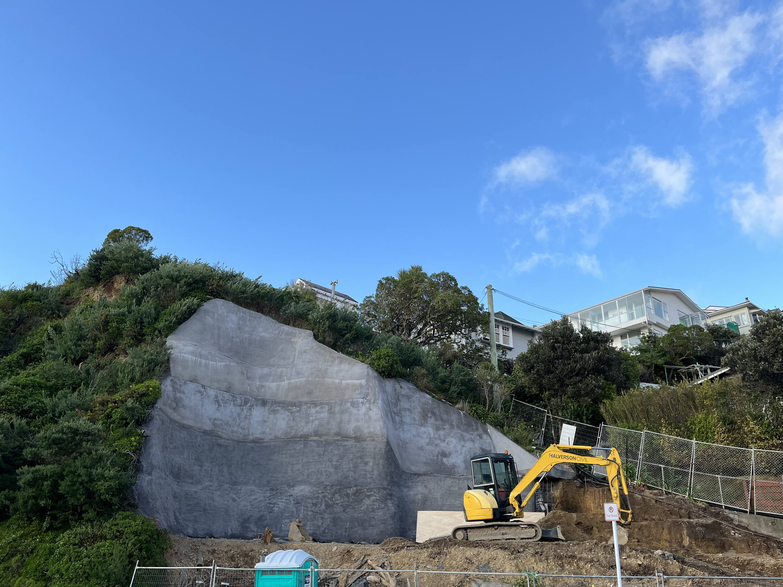 Construction site on a hillside with a small yellow excavator, a concrete retaining wall, green vegetation, and residential buildings in the background under a blue sky with some clouds.