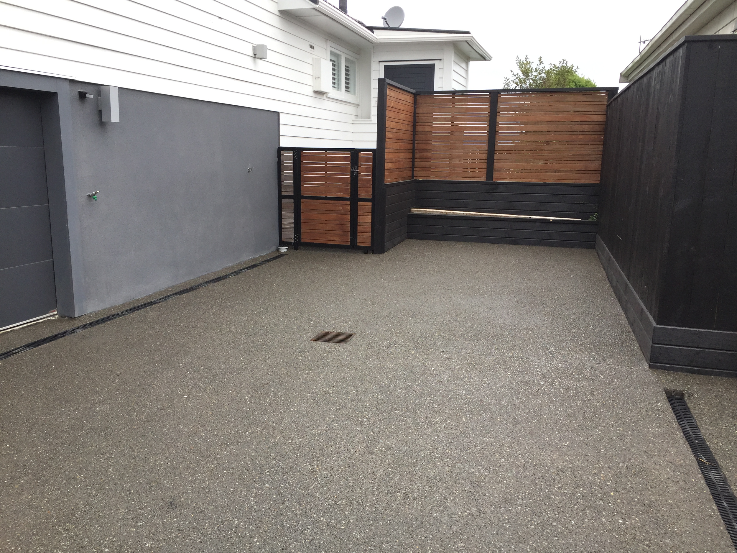 A small backyard patio area with concrete surface, fenced on three sides with a combination of wood and painted black panels, attached to a white house with horizontal siding and a small window.
