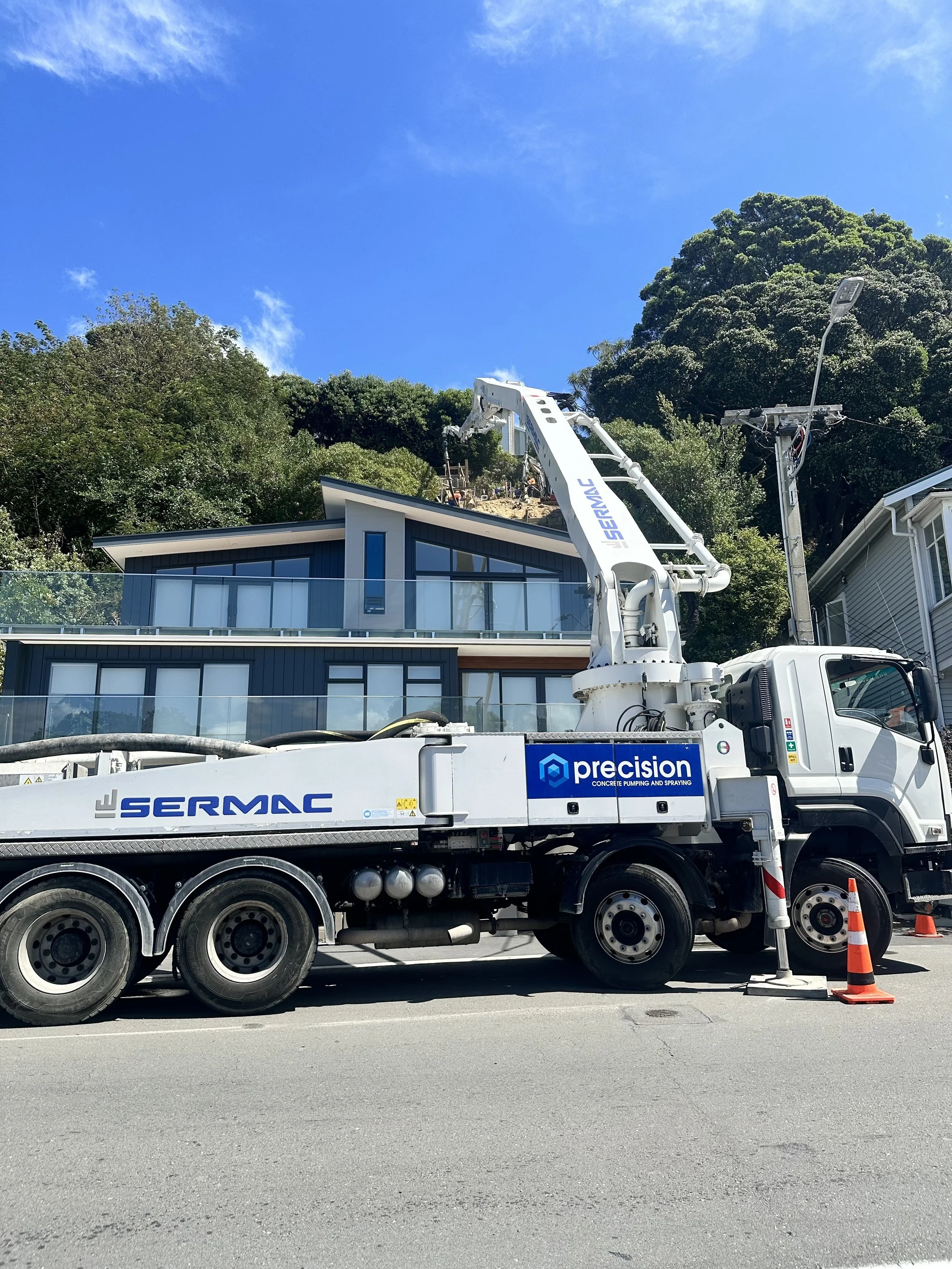 A concrete pump truck with the brand name 'Sermac' and the words 'precision concrete pumping and spraying' on the side, parked on a street with an orange traffic cone nearby. In the background, there is a house, utility poles, trees, and blue sky with some clouds.