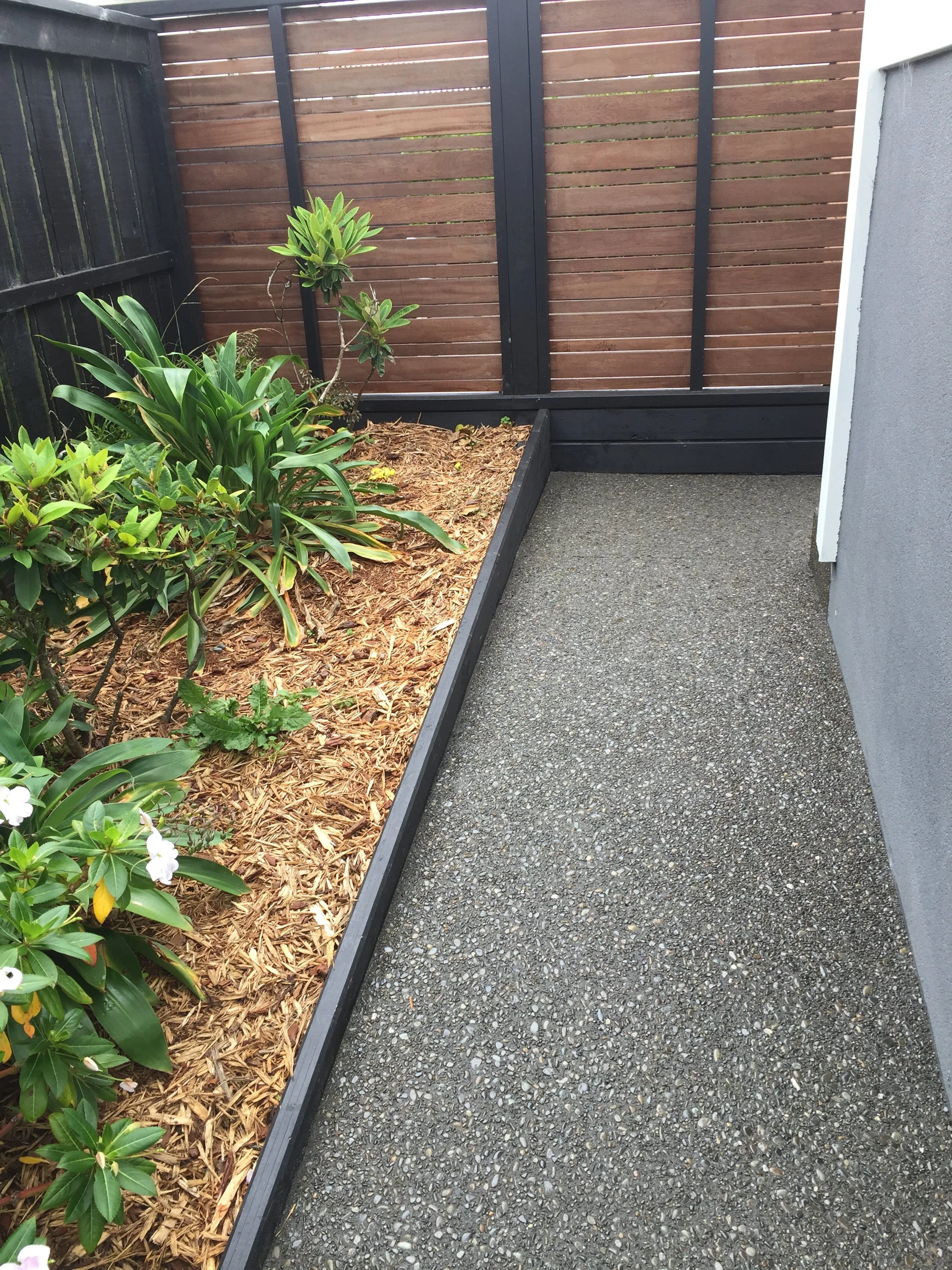 A small landscaped outdoor area with a narrow gravel pathway, a raised garden bed with green plants and mulch, and a wooden privacy fence.