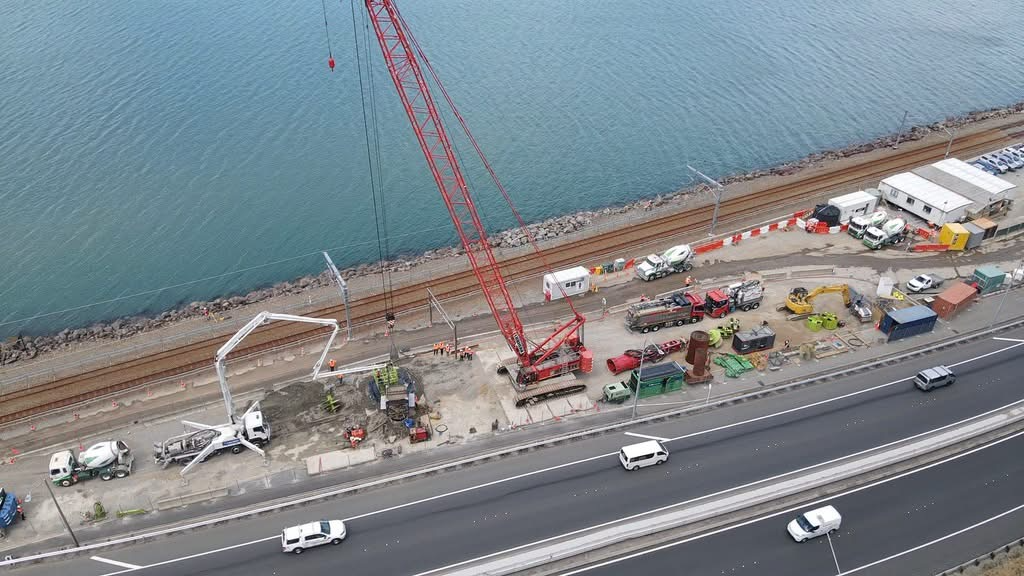 Construction site along a highway next to a body of water, with cranes, cement mixers, and construction equipment.