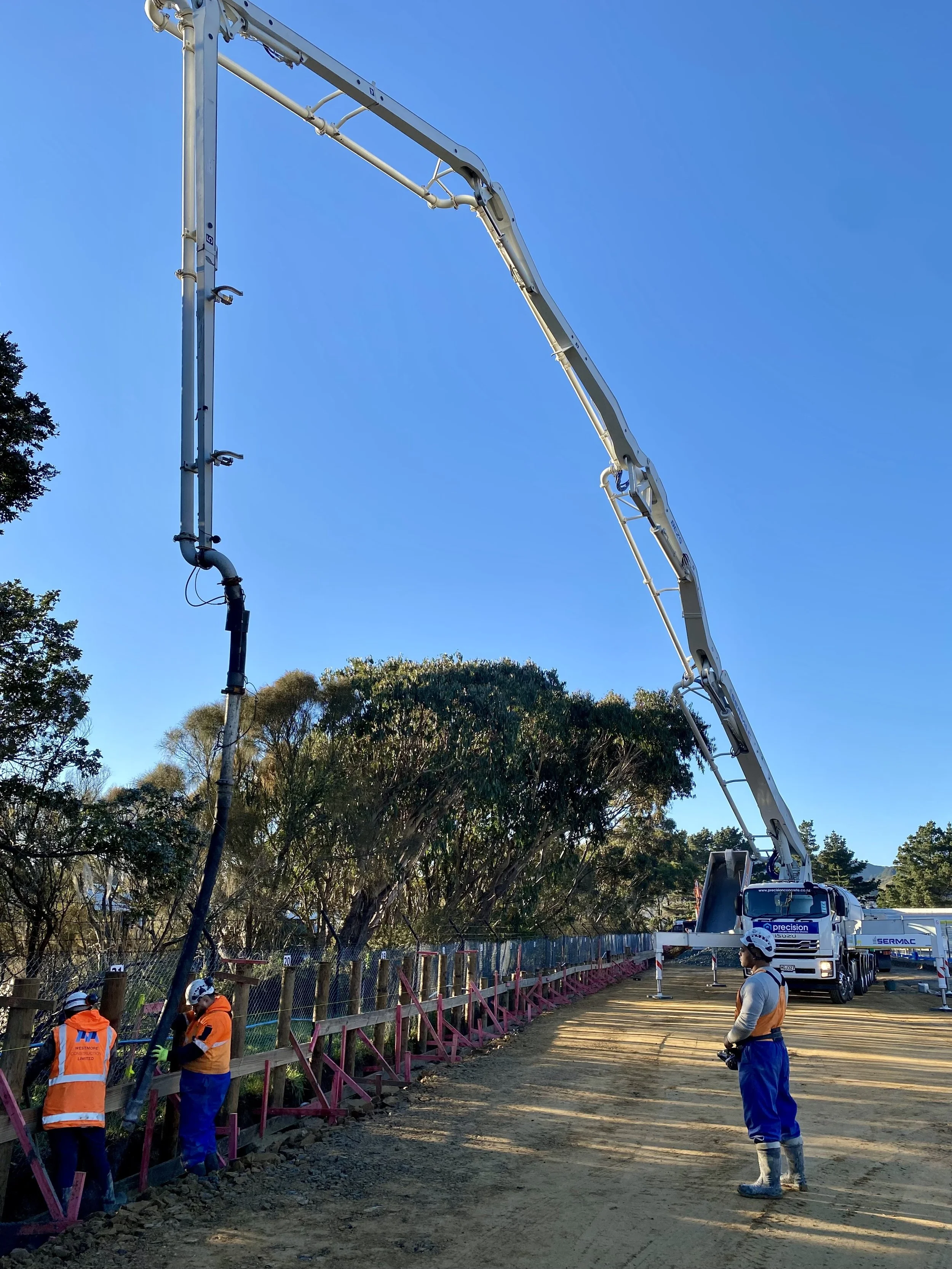 Precision Concrete Pumping and Spraying  workers in orange and blue safety gear working near a fence, with a concrete pump truck and a blue sky overhead.