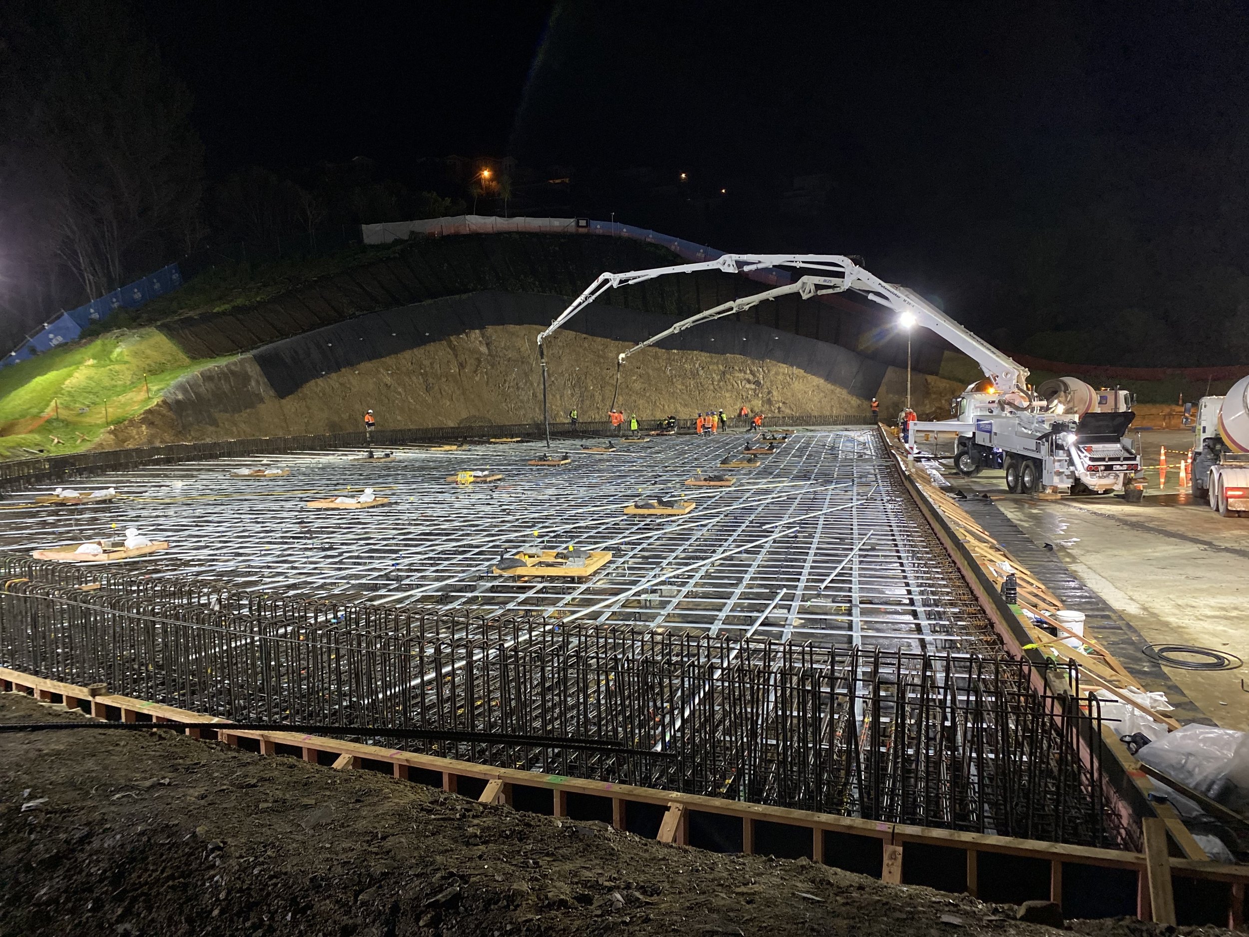 Nighttime view of a construction site with workers and a concrete pump pouring foundation, reinforced steel rebar grid in place for the building's floor, and a hillside with protective fencing.