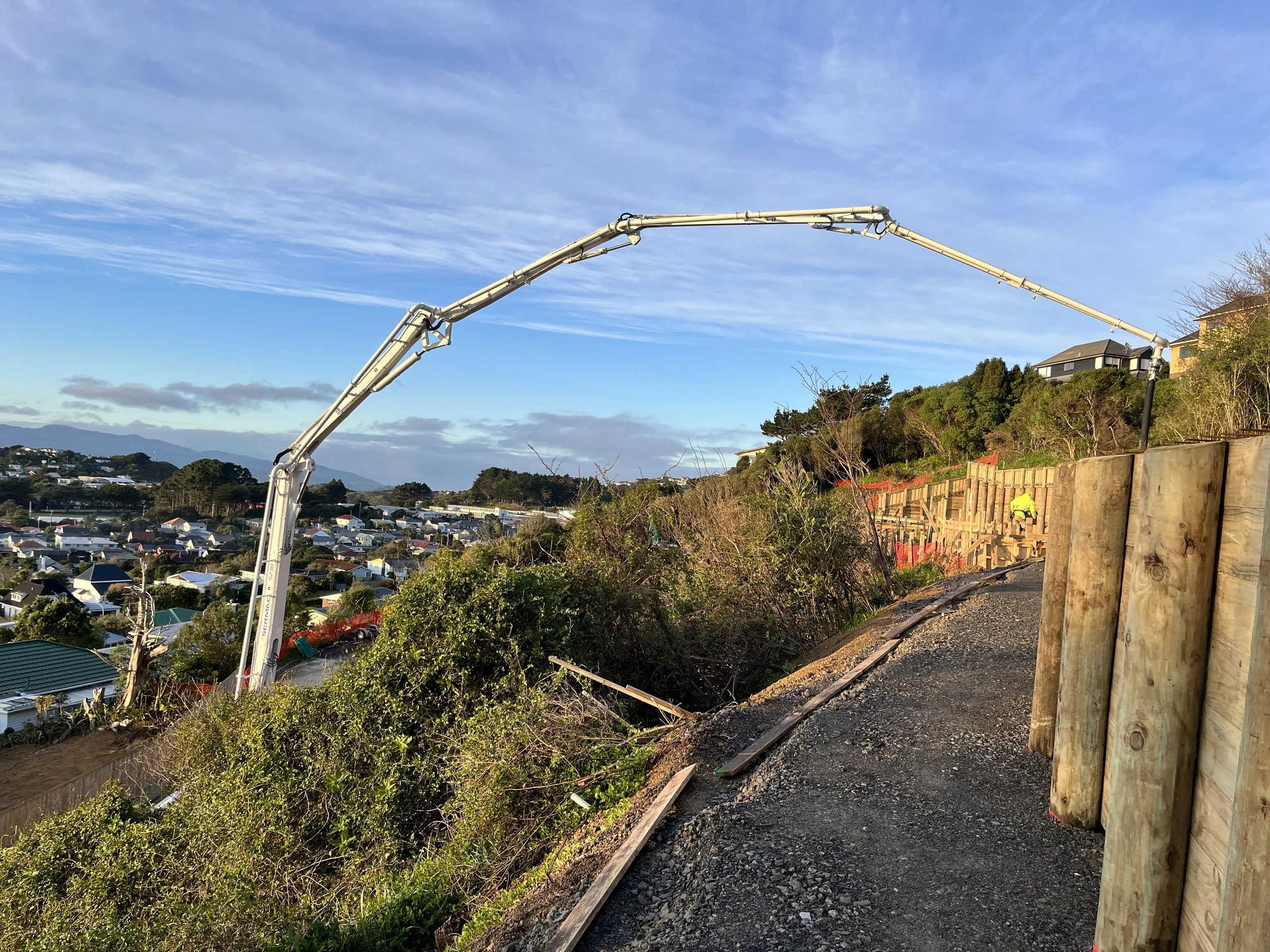 Construction site on a hillside with a concrete pump truck, a worker in a yellow safety vest, residential houses in the background, green trees, and a blue sky with clouds.