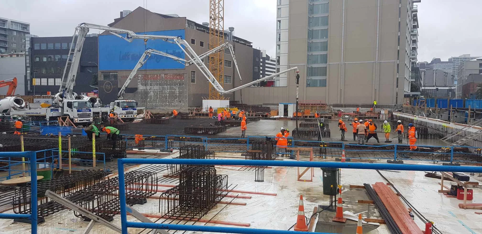 Construction workers in orange and green safety vests and helmets working on a building foundation with rebar and concrete, surrounded by construction equipment and machinery in an urban area.