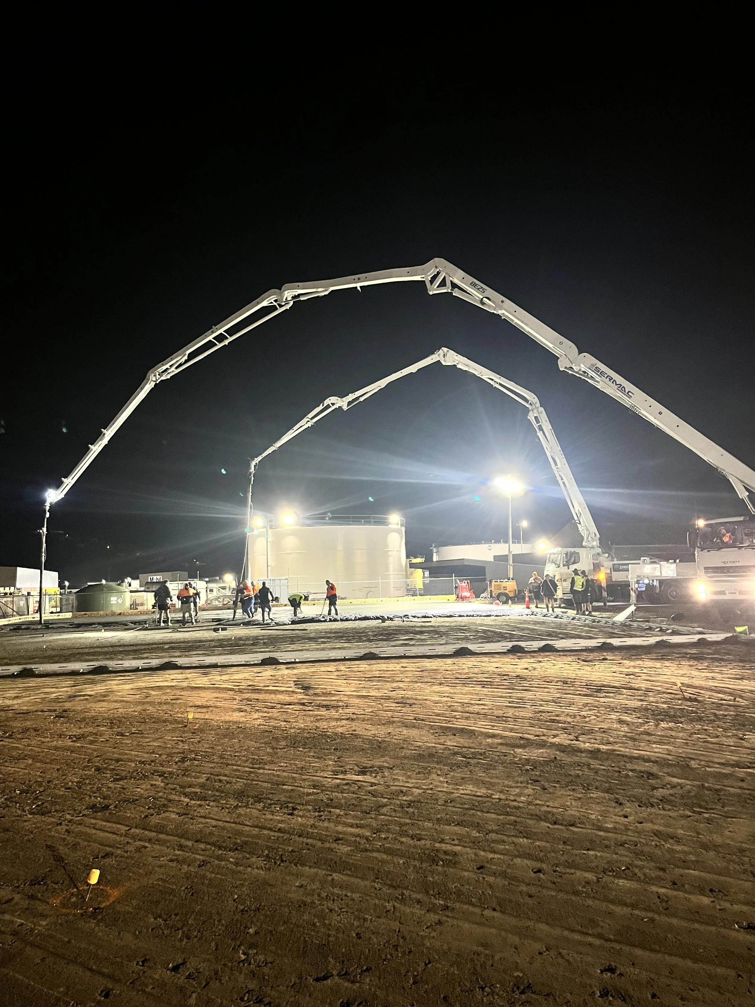 Construction site at night with concrete pump trucks, workers, and equipment illuminated by bright lighting against a dark sky.