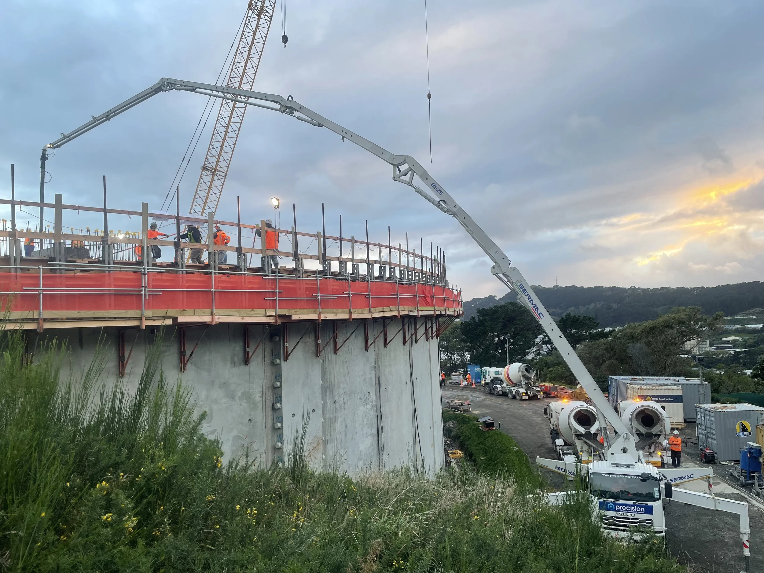 Construction workers on a building site with a concrete pump truck and a crane, with a hilly landscape and cloudy sky in the background.