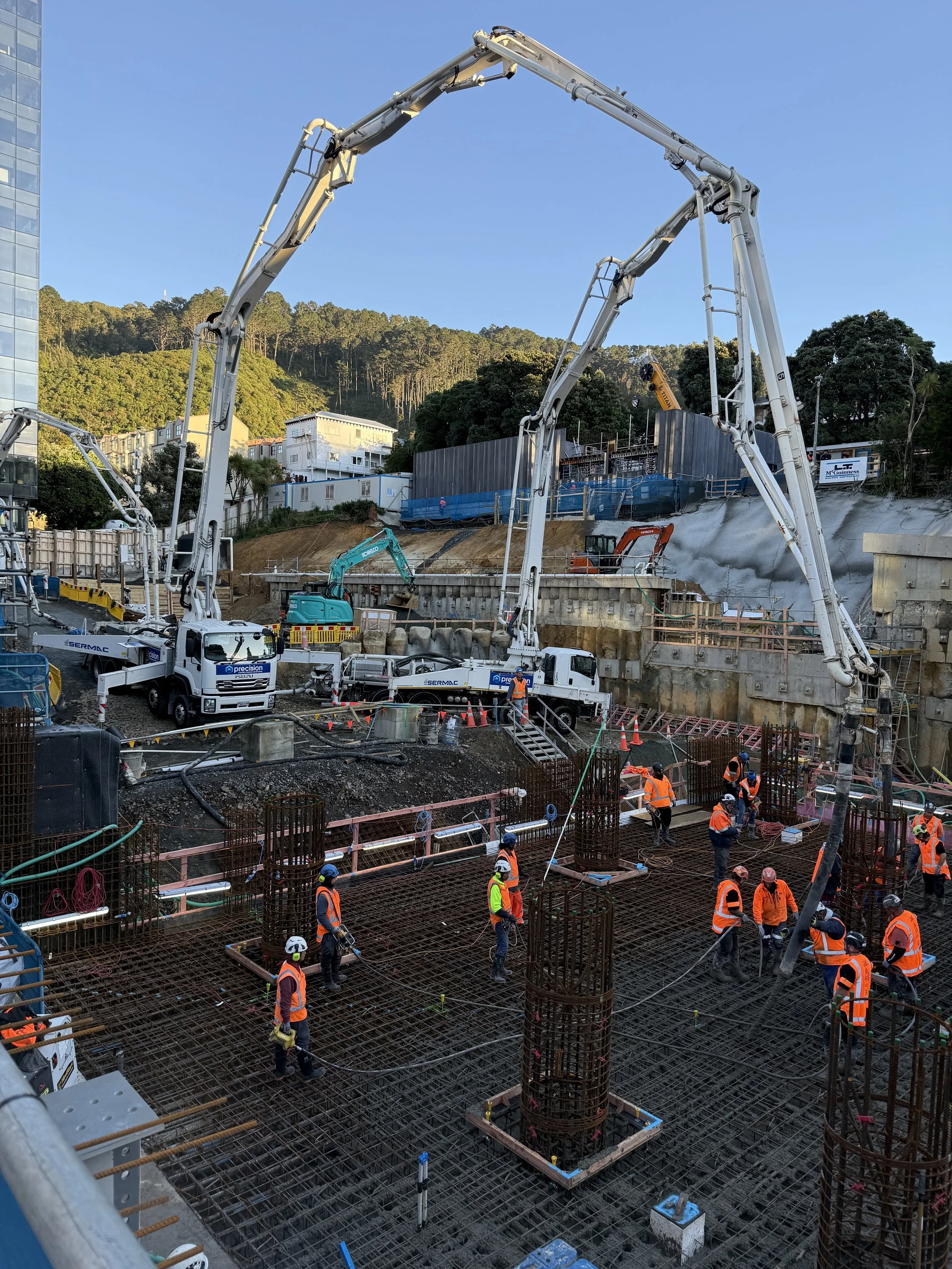 Construction workers wearing safety vests and helmets working on a building site with rebar and concrete foundation, using large machinery under clear blue sky, with hills and buildings in background.