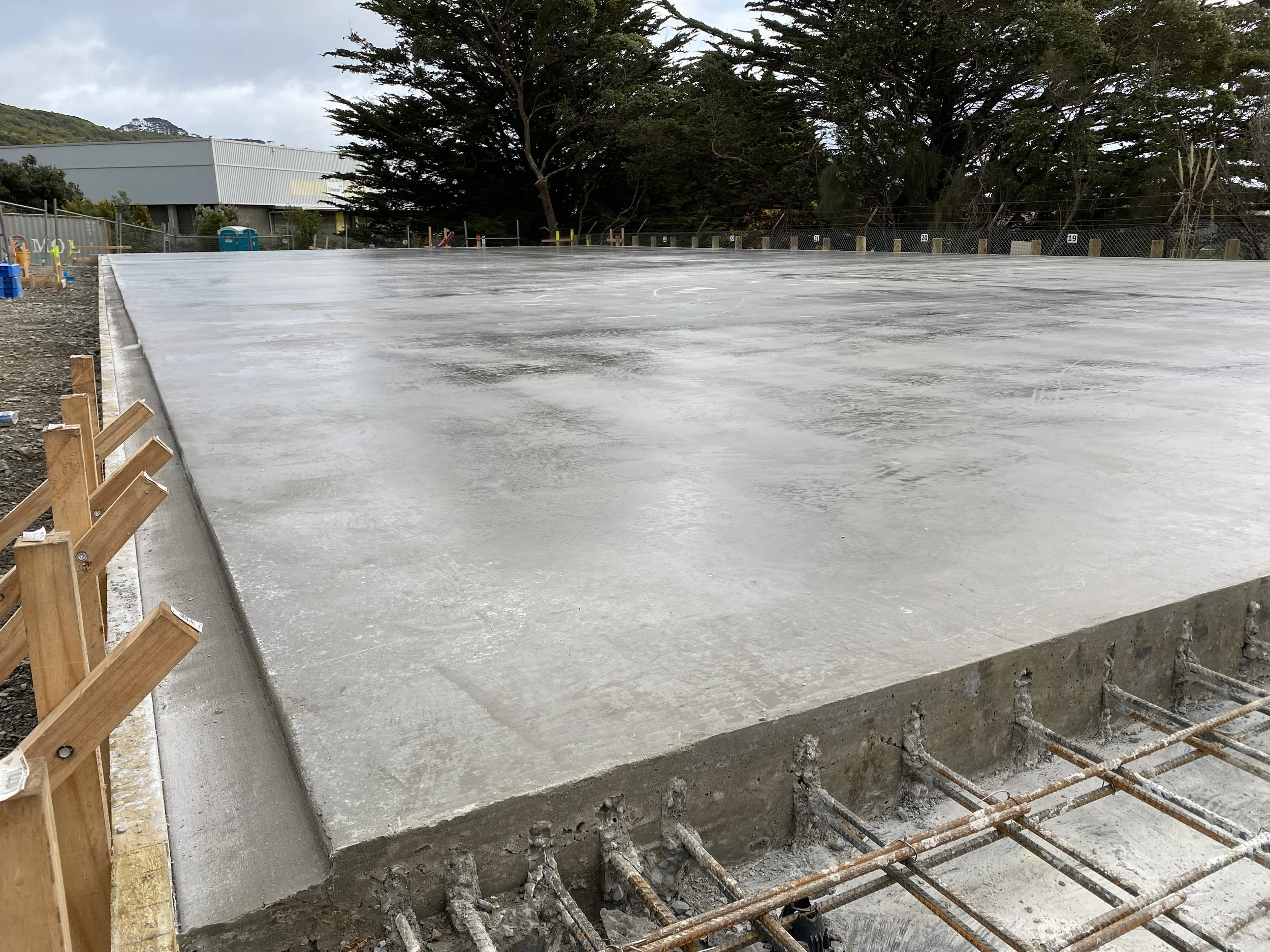 Concrete foundation slab being poured on a construction site with wooden formwork and rebar, surrounded by trees and a fence.