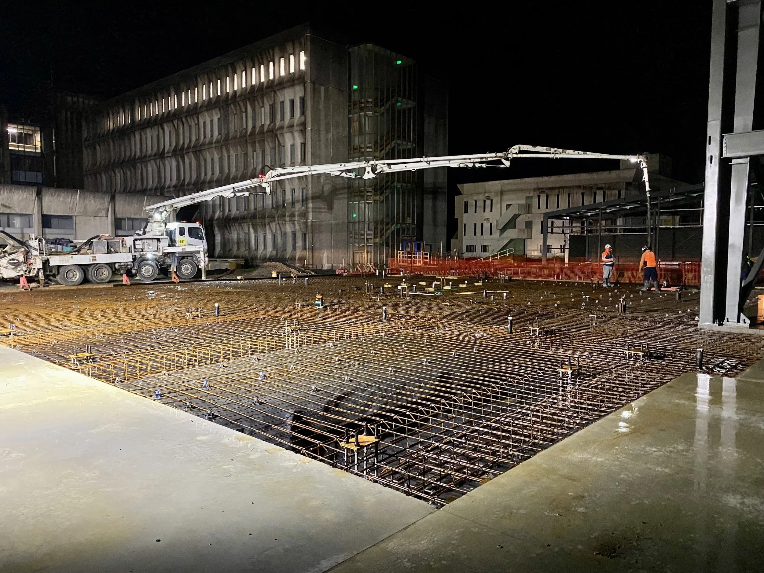 Nighttime construction site featuring steel reinforcement grid with two workers discussing, a concrete pump truck in the background, and a modern building with parking structure and green lights.
