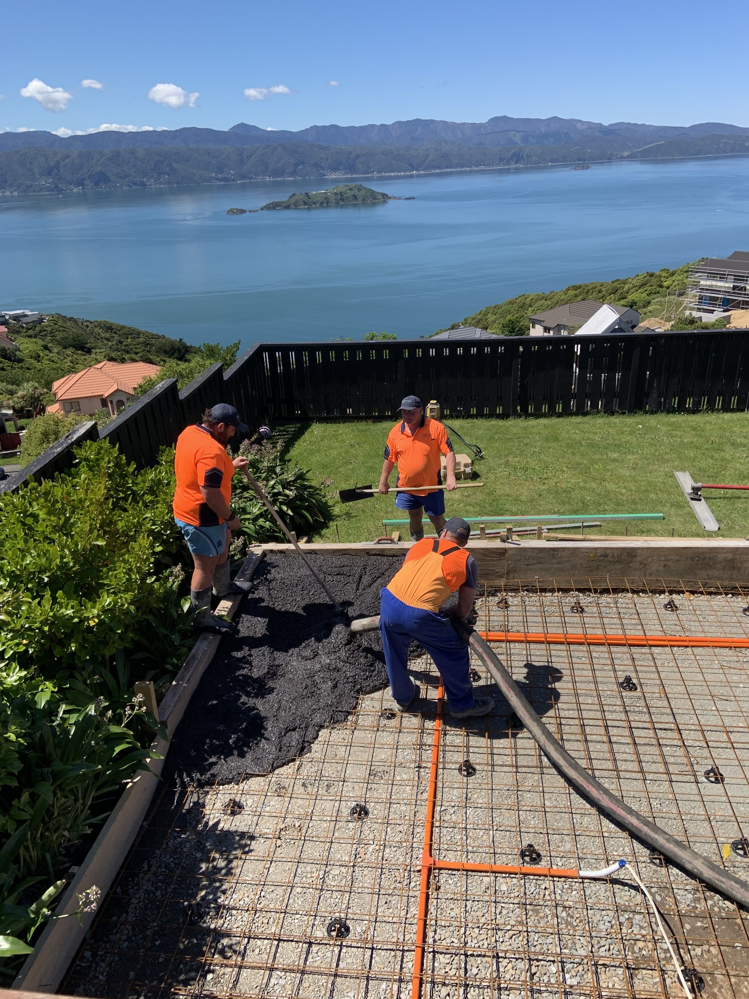 Workers pouring concrete on a building foundation with a scenic view of a large body of water, islands, and mountains in the background.