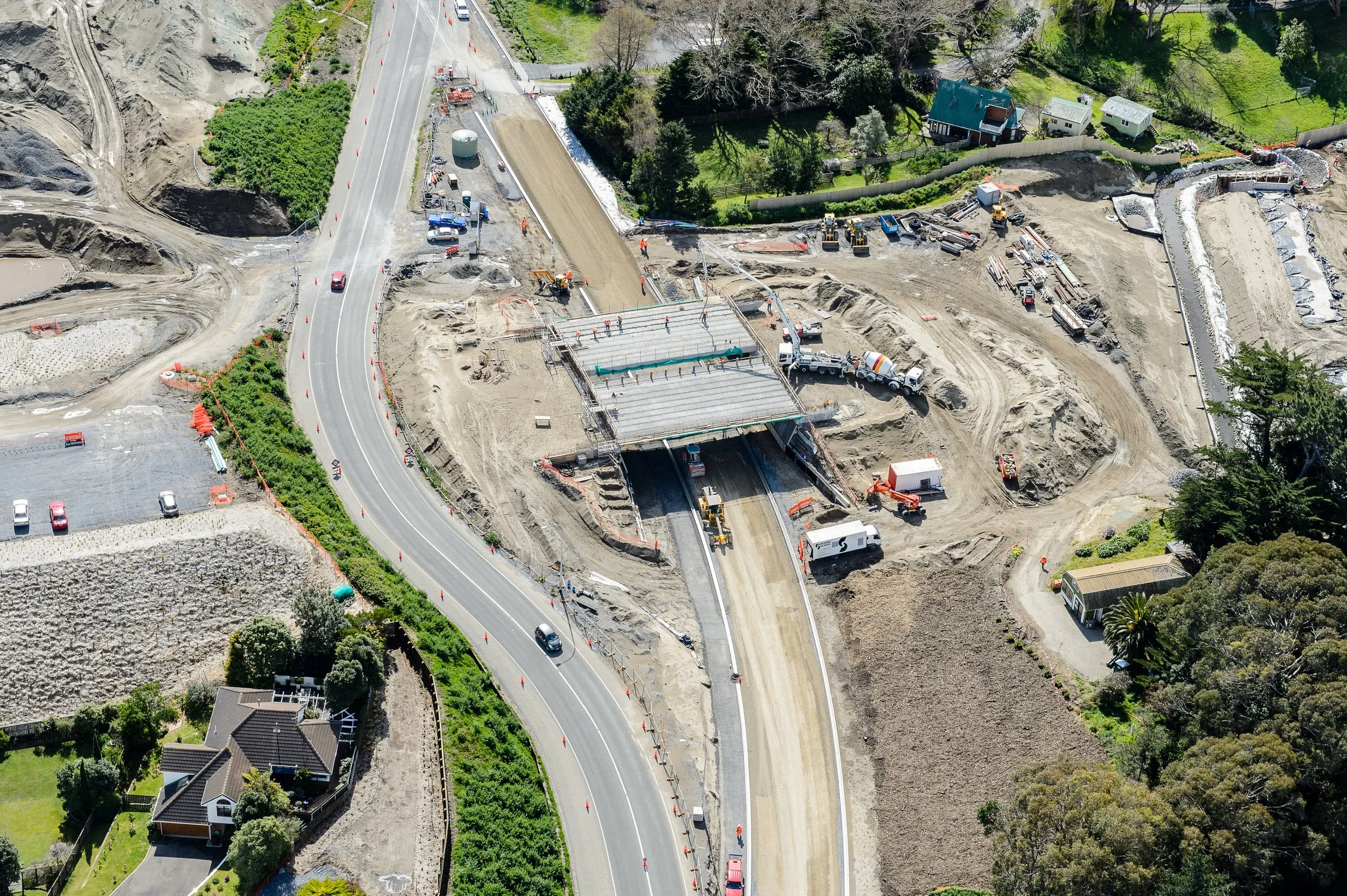 Aerial view of a construction site on a hillside with ongoing roadwork and excavation, surrounded by residential houses and green trees.