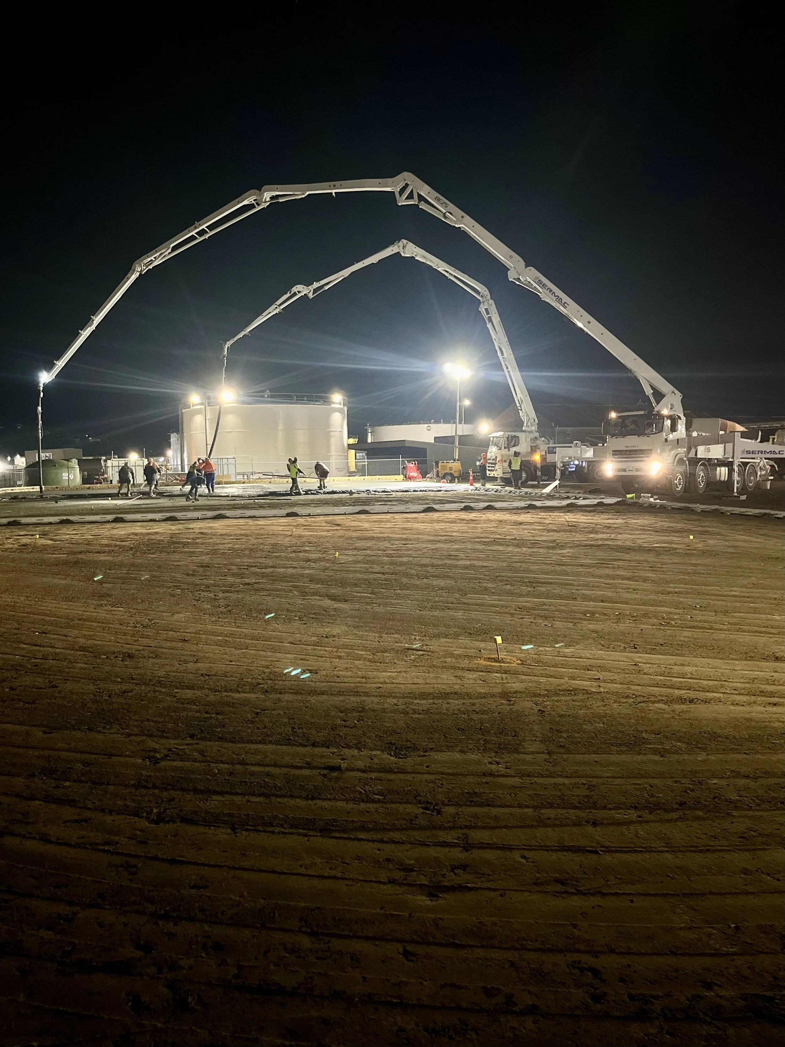 Construction site at night with multiple concrete pump trucks pouring concrete into large tanks, workers on site, surrounded by fencing and illuminated by bright lights.